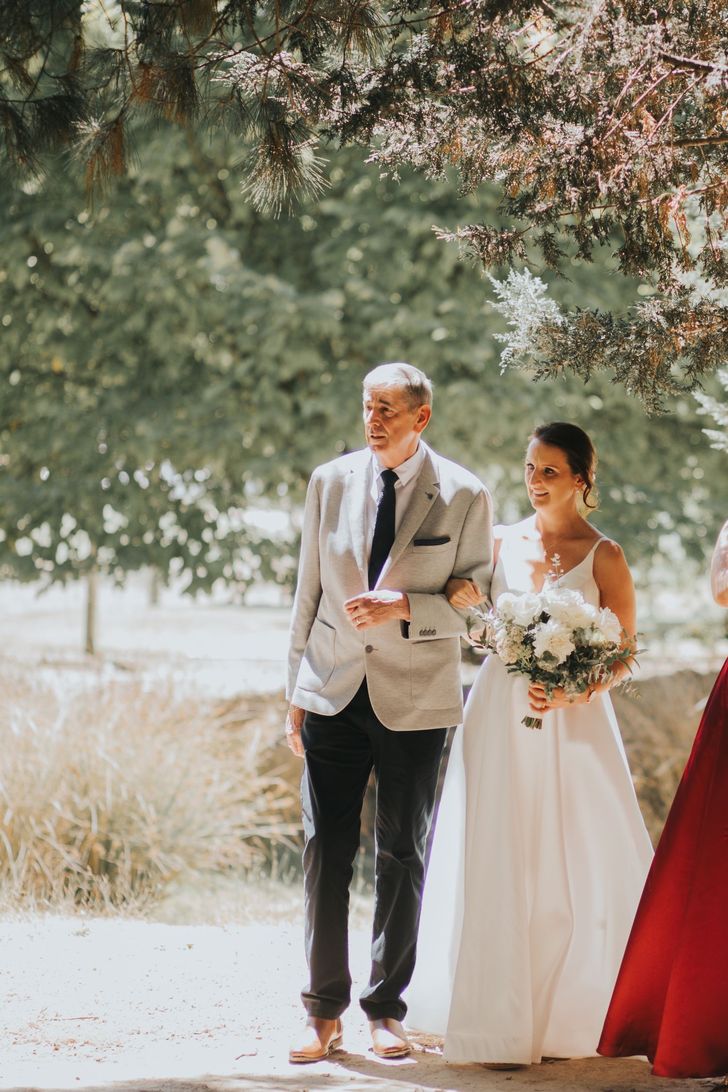 A smiling bride in a white gown holding a bouquet walks down the aisle with her father.