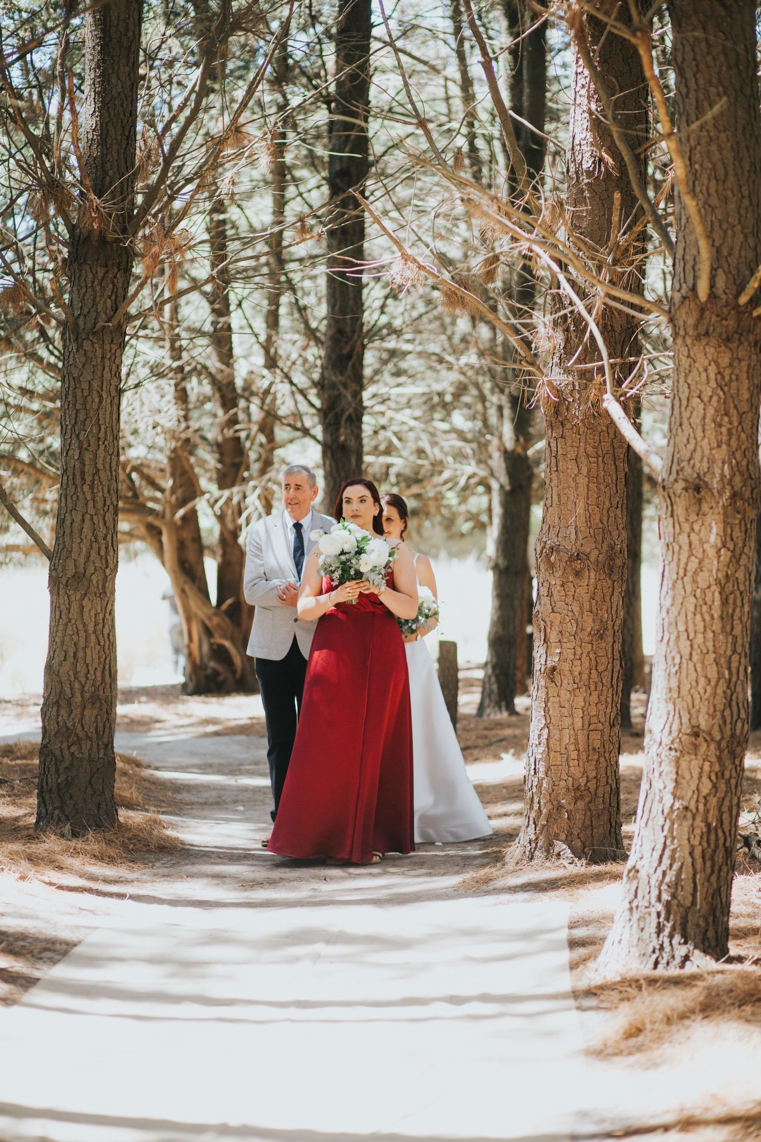 A bride in white and bridesmaid in red walk through a sunlit pine tree lined wedding aisle.
