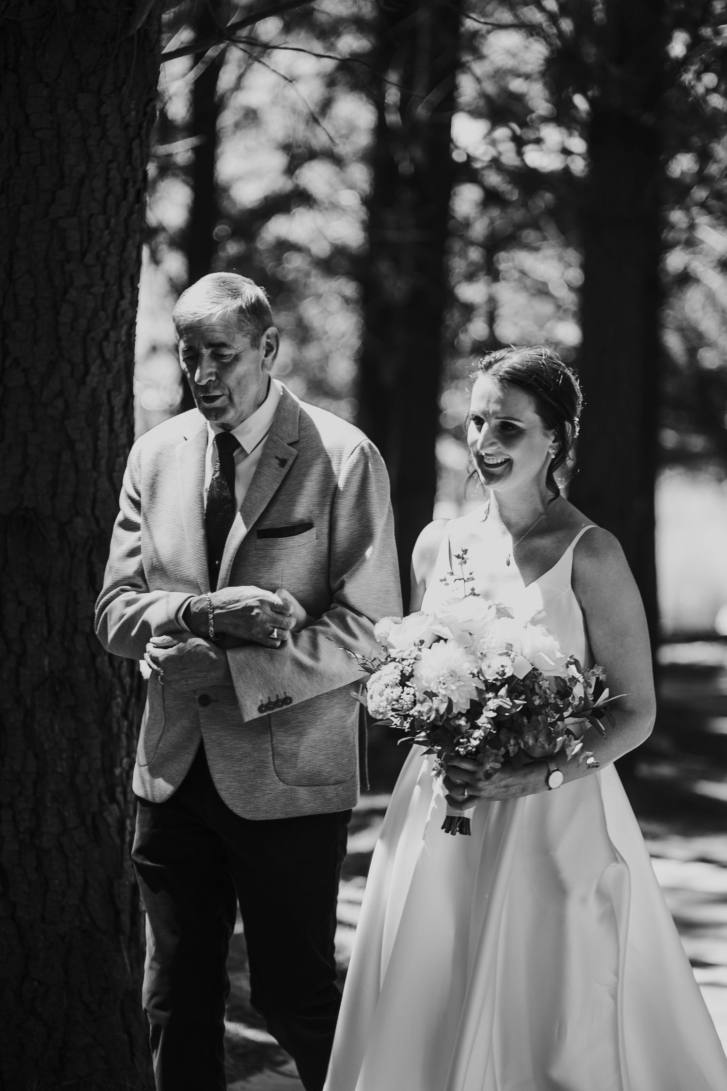 Black and white image of a laughing bride holding a bouquet walking with her father outdoors.