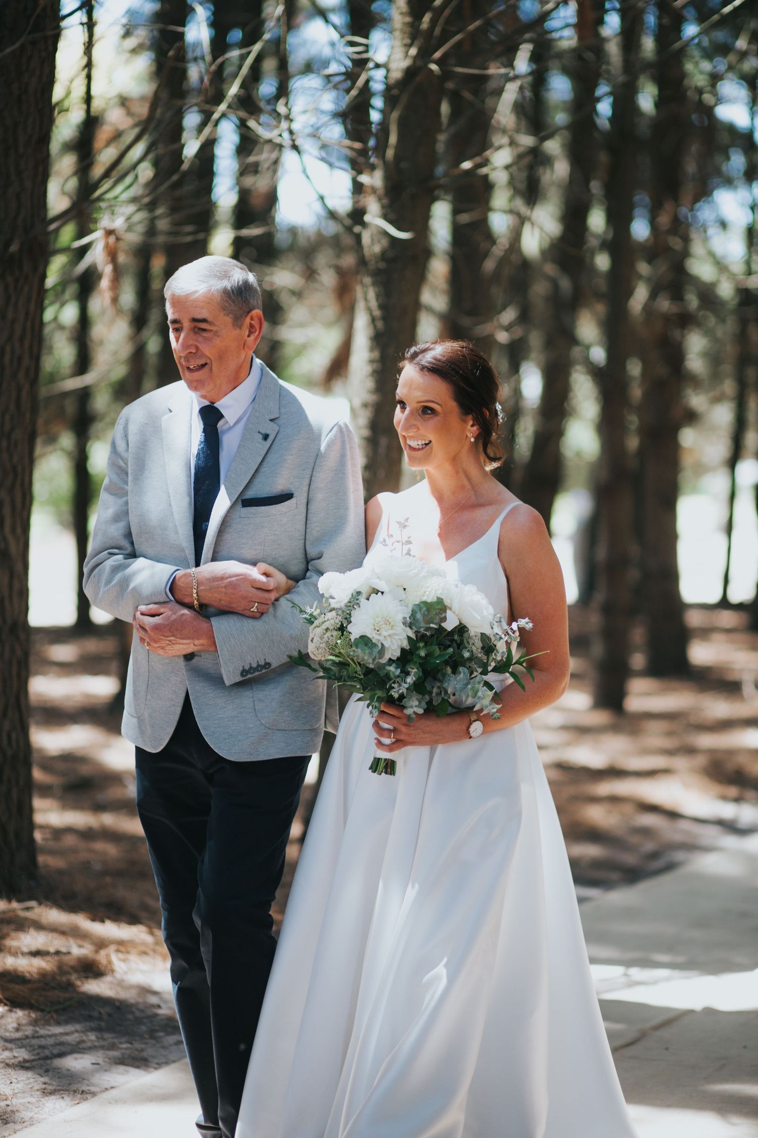 A radiant bride in a white gown walks with her father through a pine tree lined aisle.