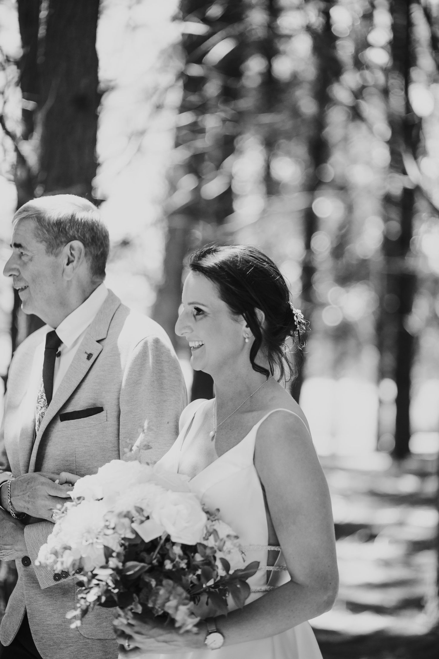 Black and white photo of a smiling bride holding a bouquet walking with her father outdoors.