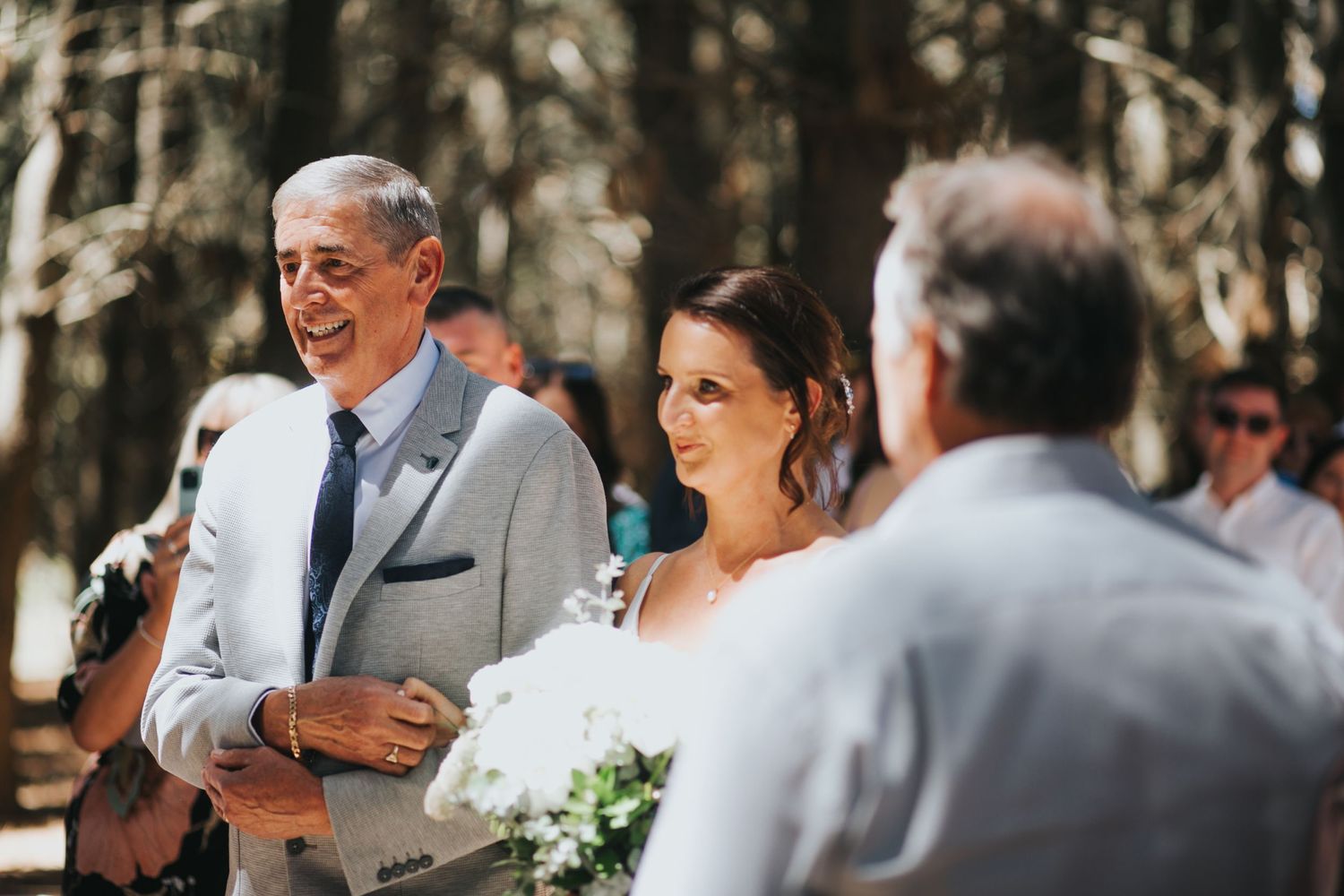 A bride and her father arrive at the altar smiling during an outdoor woodland wedding ceremony.