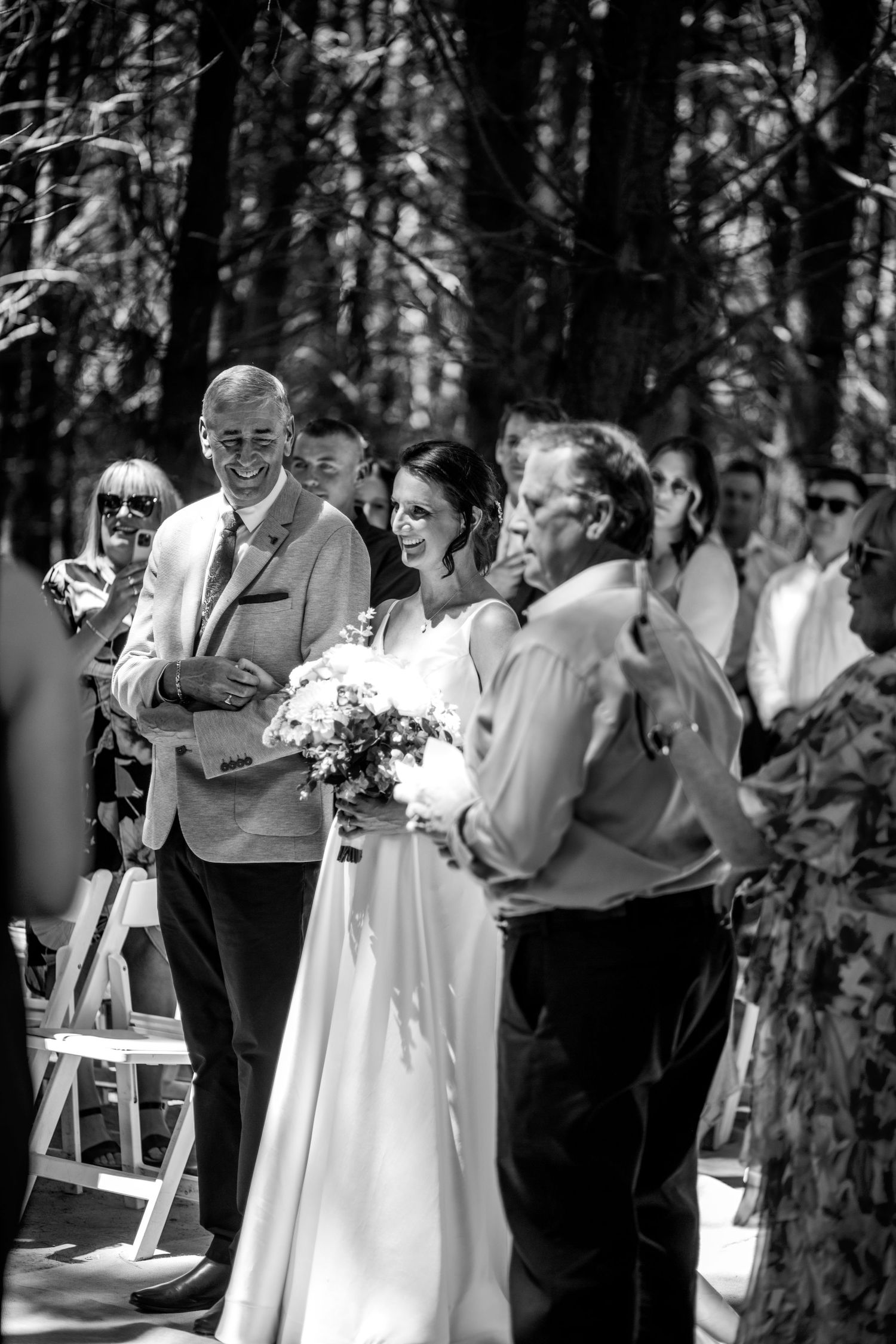 Bride walks down forest aisle smiling, holding bouquet, escorted by two men at outdoor wedding ceremony.