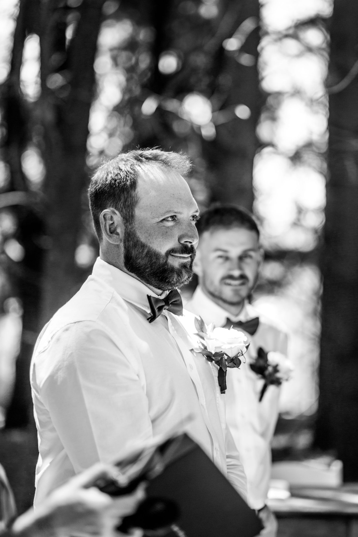 Bearded groom in white shirt and bow tie watches bride approach at outdoor forest wedding ceremony.