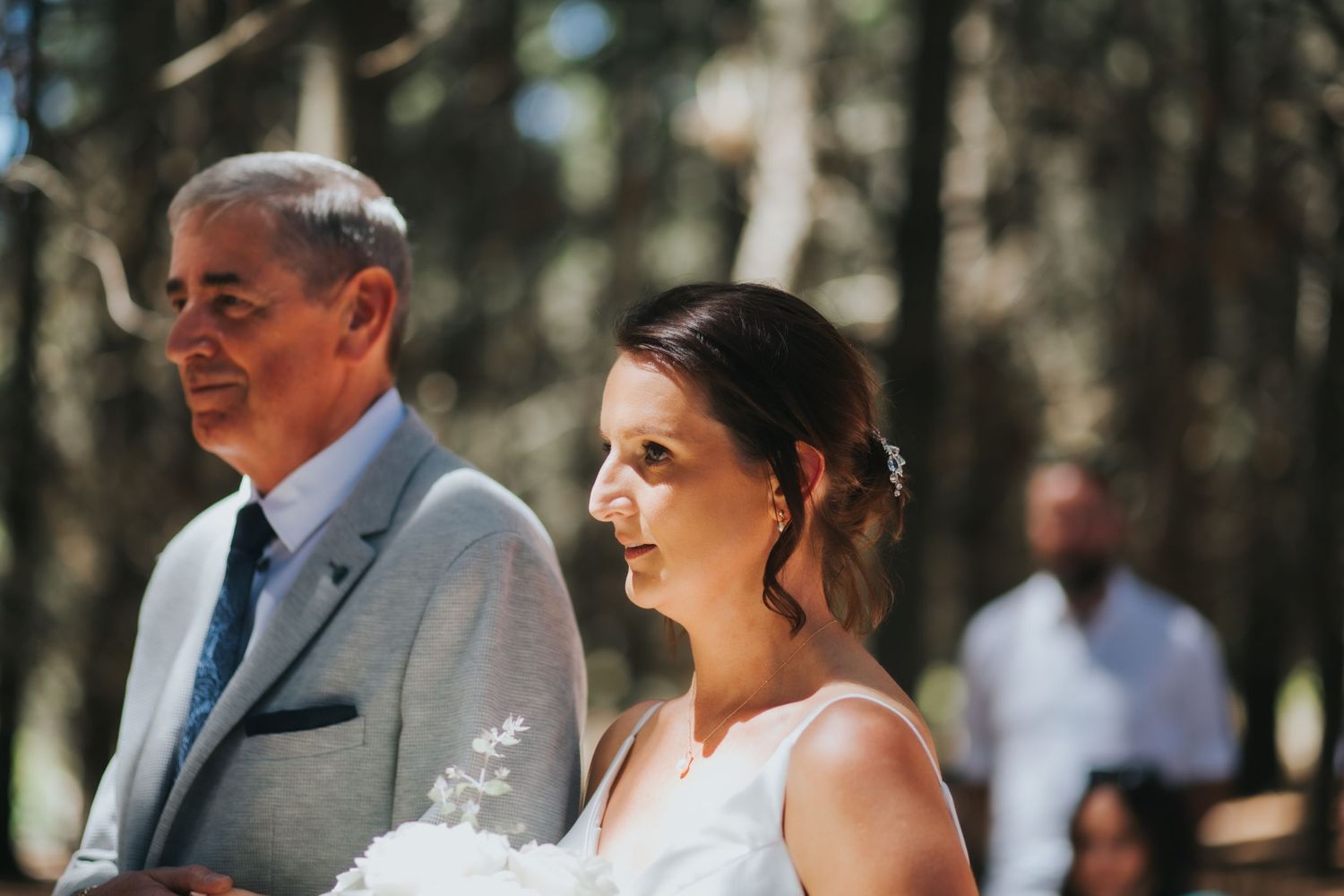 Bride holds white bouquet walking down aisle with older man in grey suit at forest wedding.