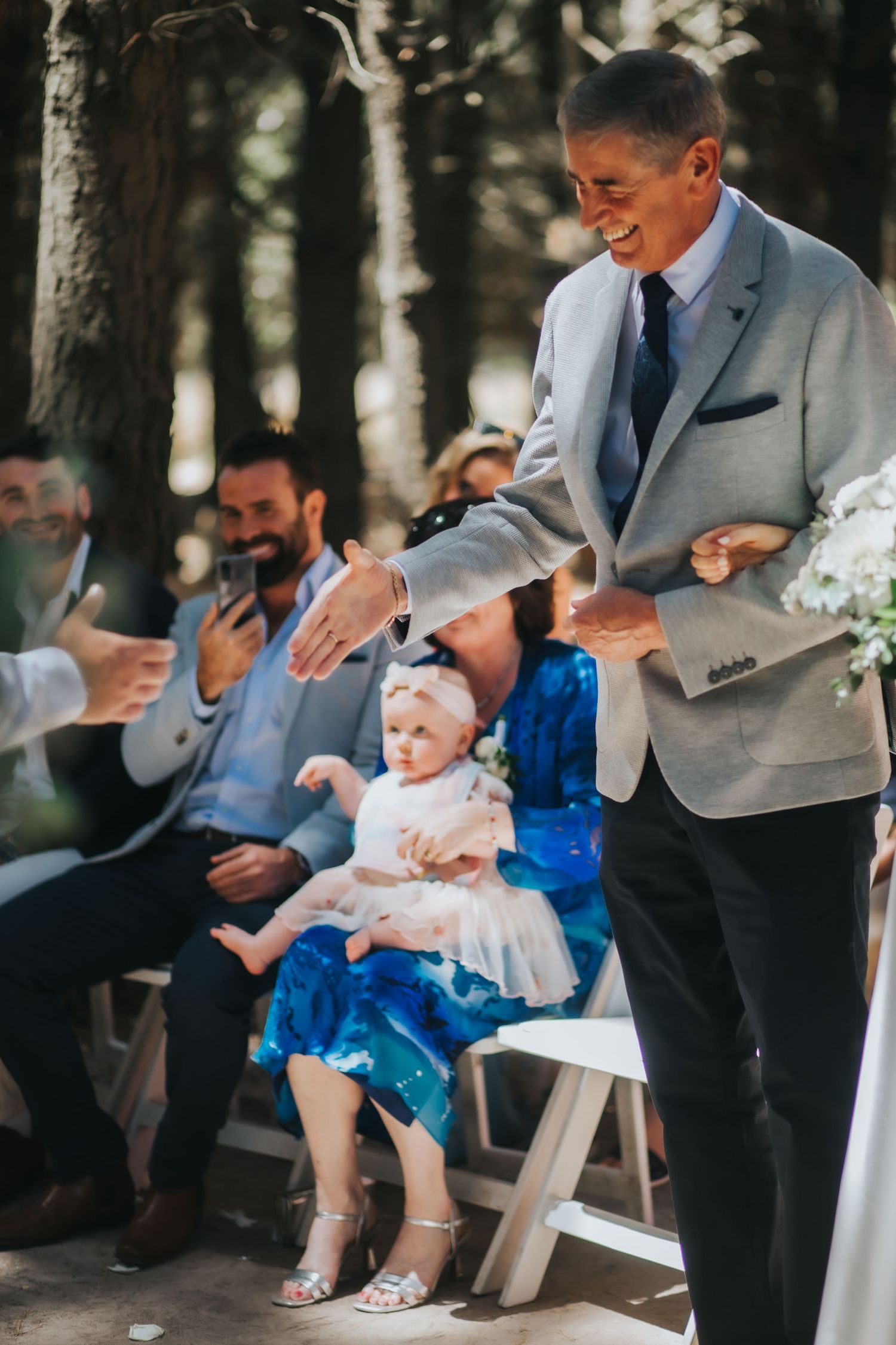 Smiling man in grey suit greets seated guests including baby at outdoor forest wedding ceremony.