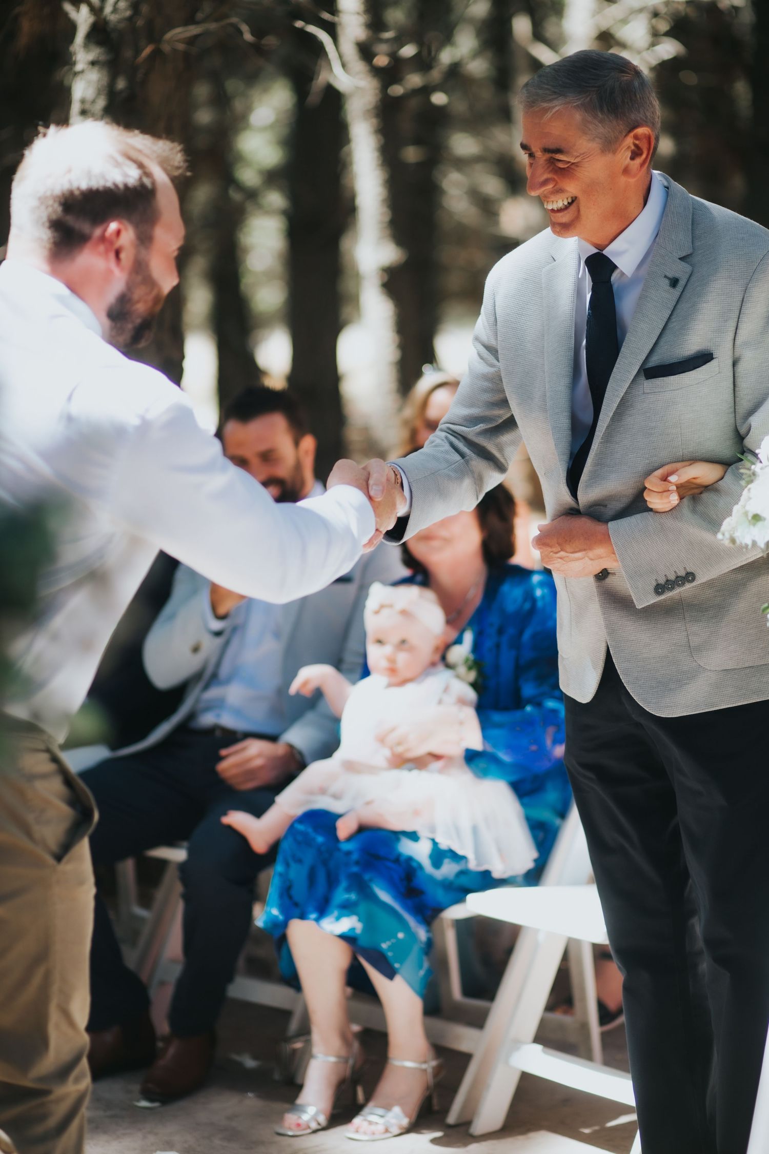Two men share a handshake and smile at outdoor forest wedding ceremony with seated guests behind.