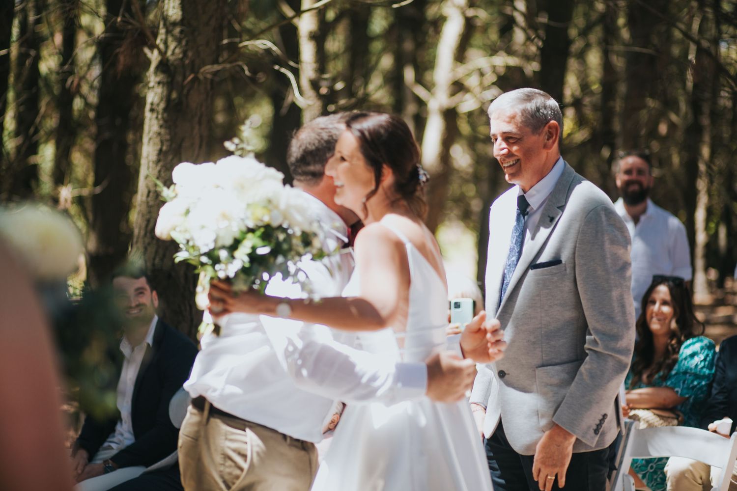Bride kisses guest while man in grey suit smiles at sunny outdoor forest wedding ceremony.