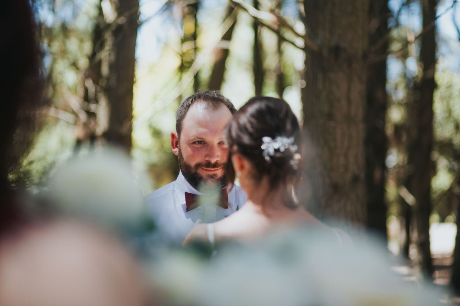 Bearded groom smiles tenderly at bride through soft bokeh blur at outdoor forest wedding ceremony.