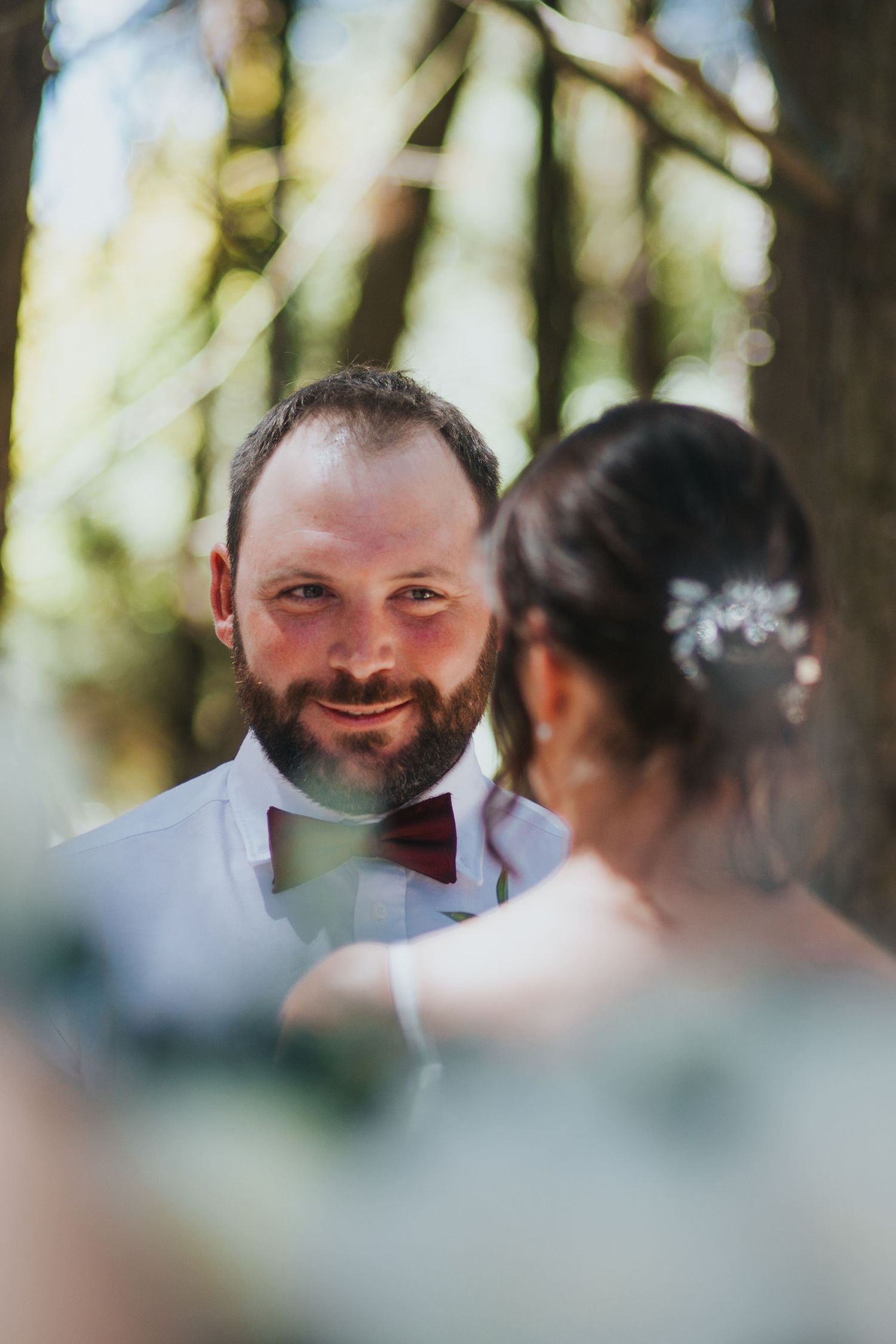 Bearded groom in white shirt and burgundy bow tie smiles at bride during forest wedding ceremony.