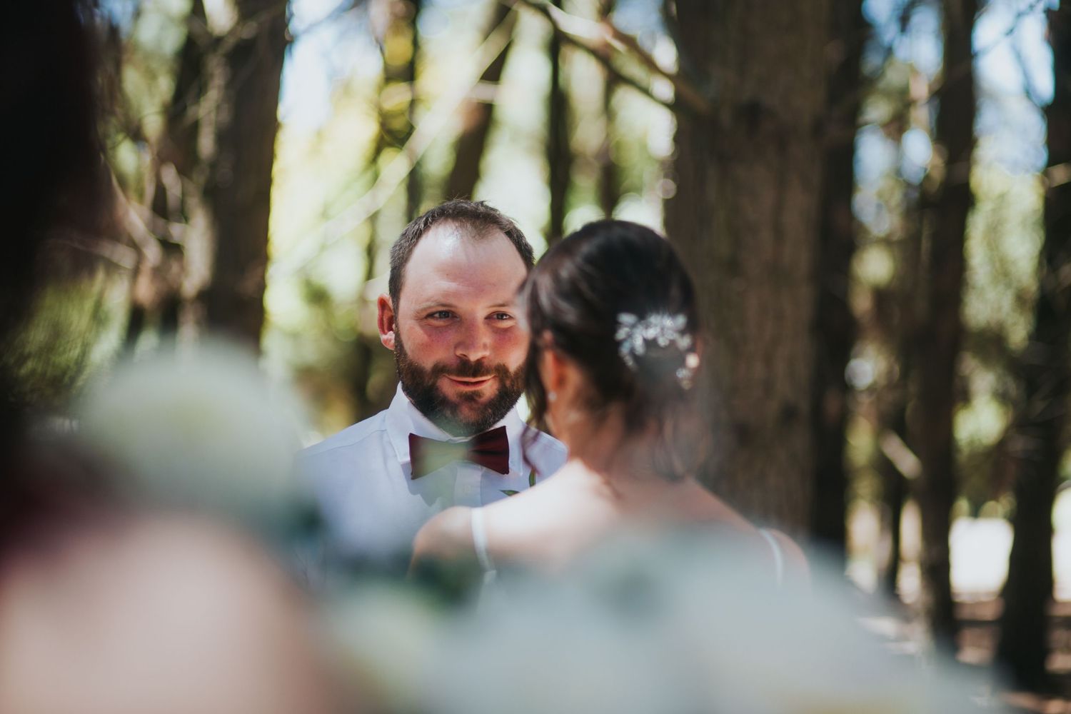 Groom with beard smiles lovingly at bride during outdoor wedding ceremony in sunlit forest.