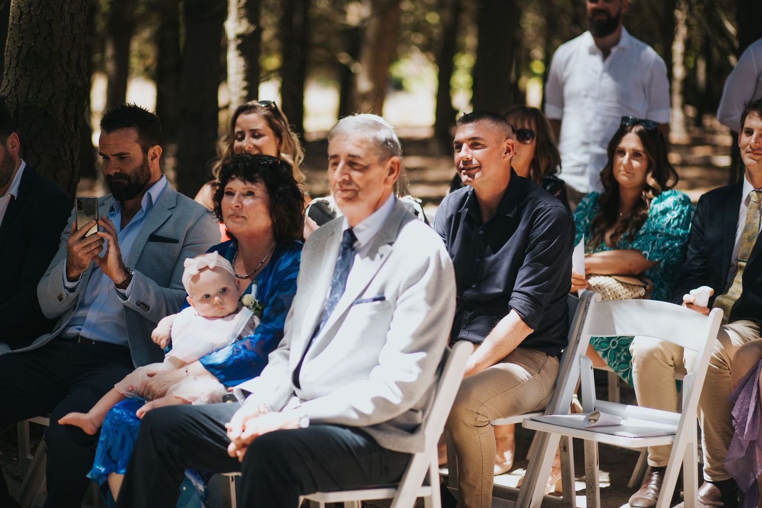 Wedding guests smile and laugh while seated outdoors, with woman holding baby in forest setting.