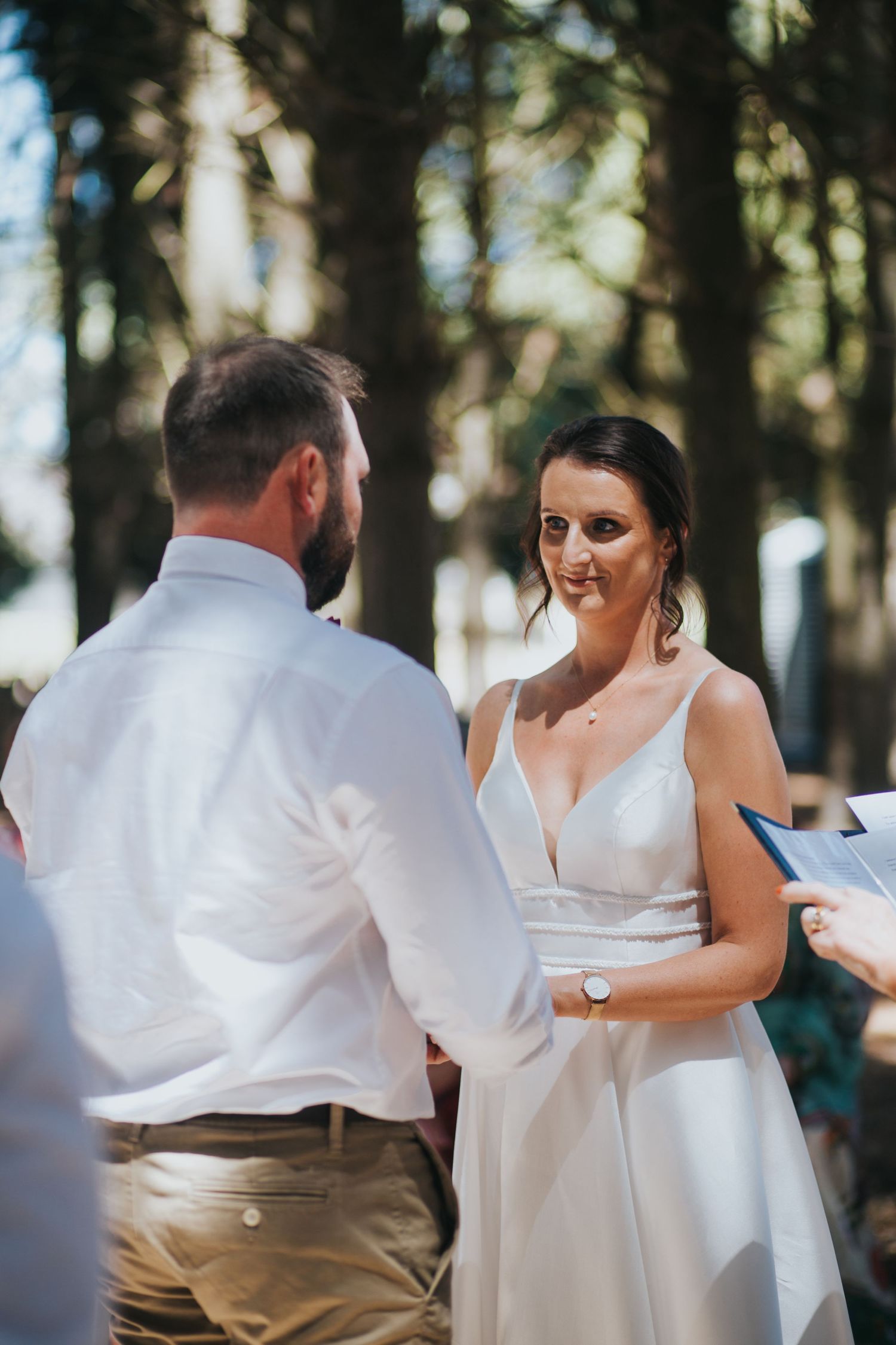 Bride in white gown smiles at bearded groom while exchanging vows at outdoor forest wedding.