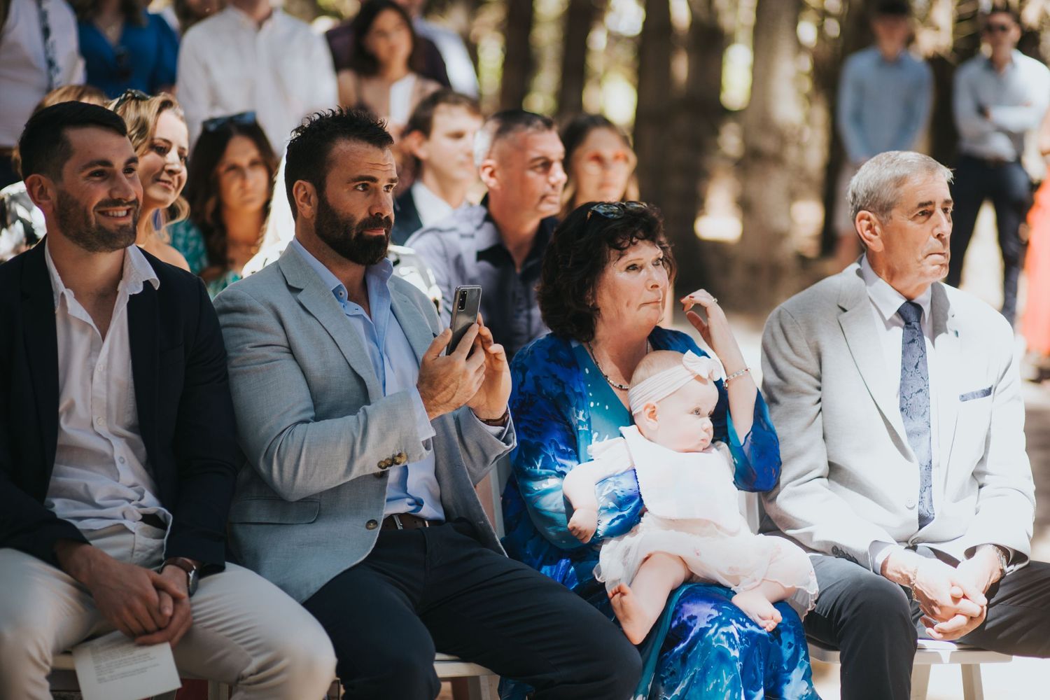 Wedding guests seated outdoors, including woman holding infant, watching ceremony in wooded venue.