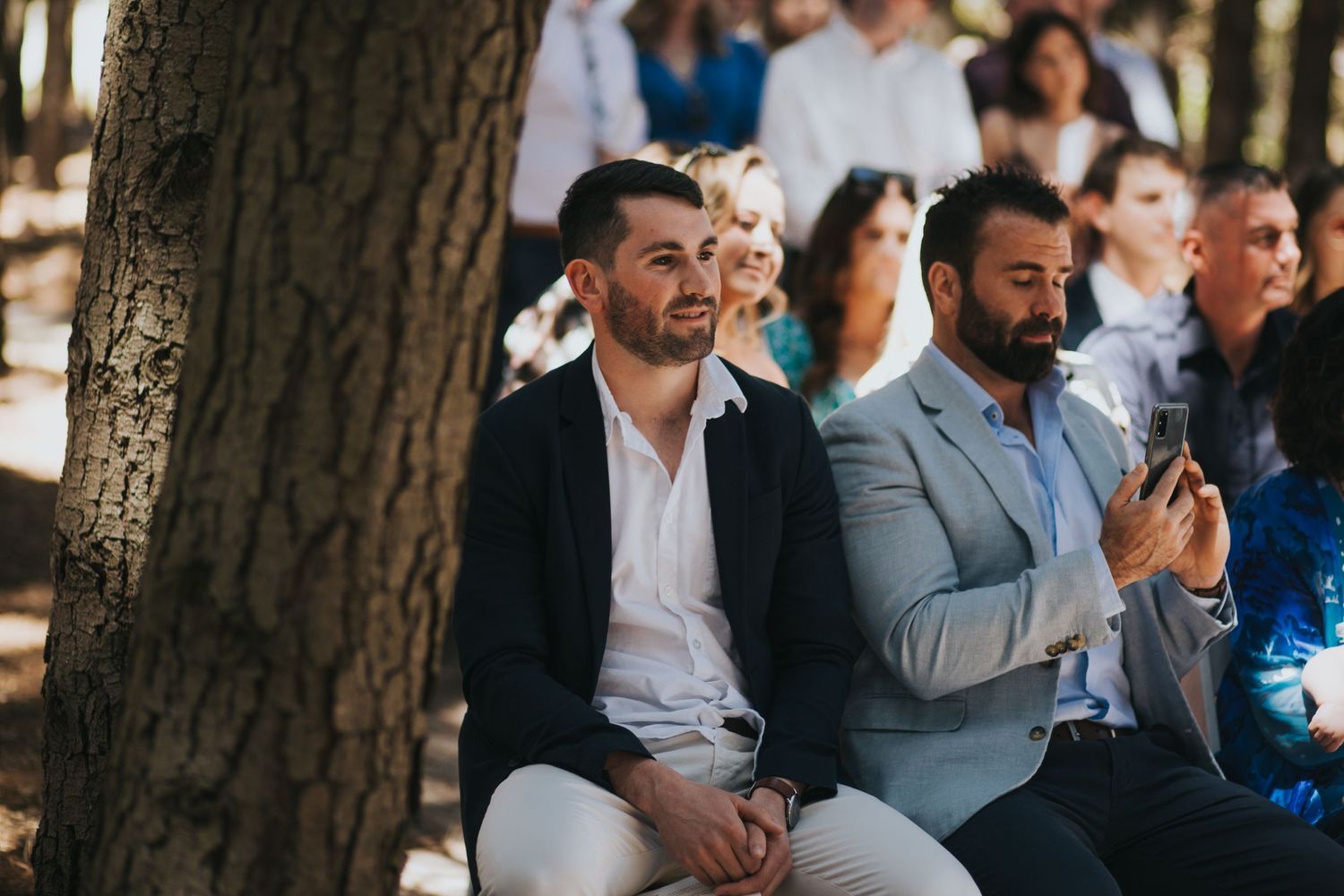 Two smiling male wedding guests seated by tree trunk enjoying outdoor forest wedding ceremony.