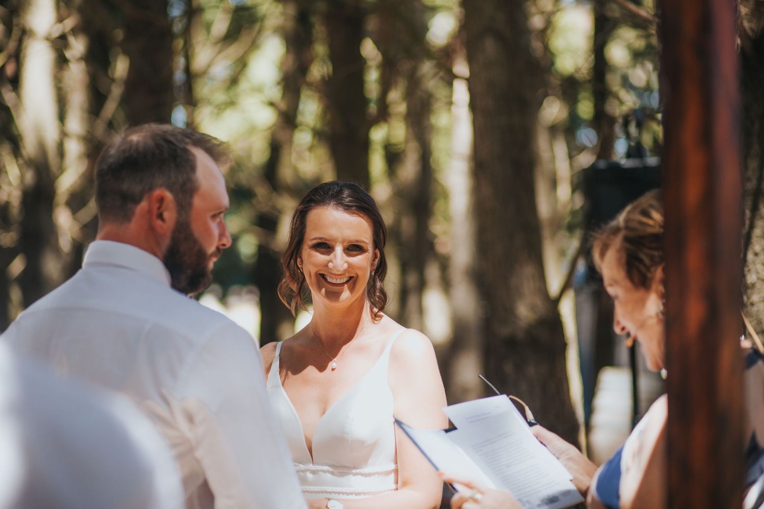 Bride smiles warmly at groom during intimate outdoor wedding ceremony in sunlit forest setting.
