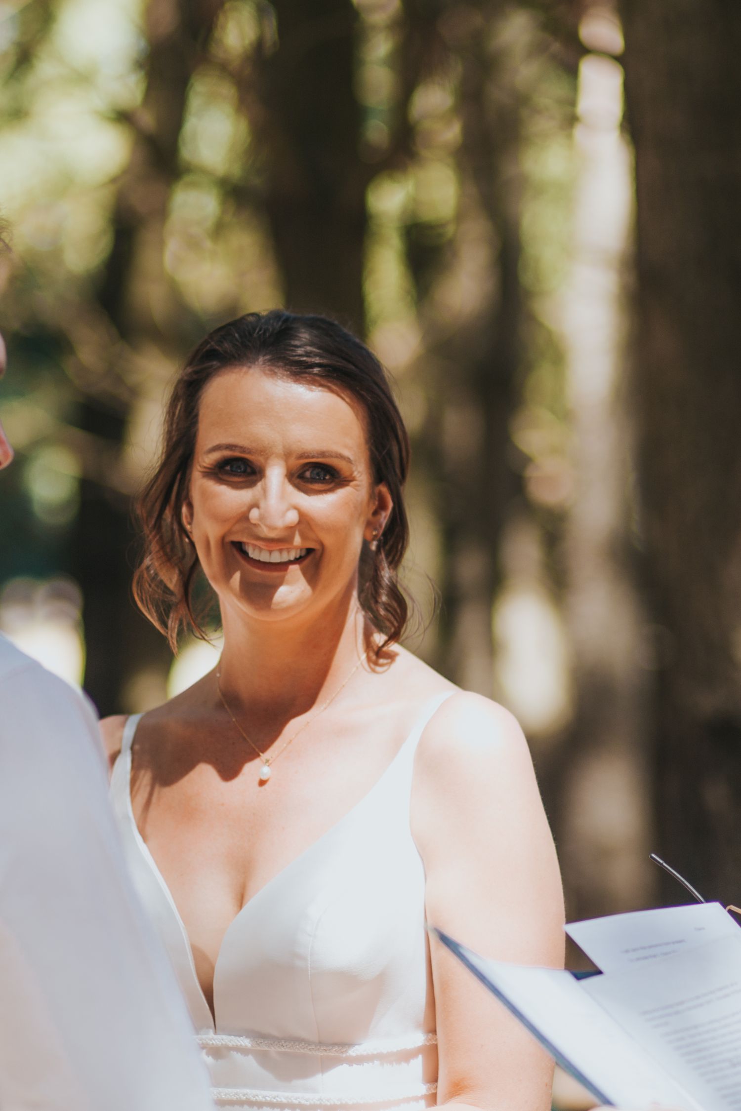Radiant bride in white gown smiles joyfully during outdoor wedding ceremony among tall trees.