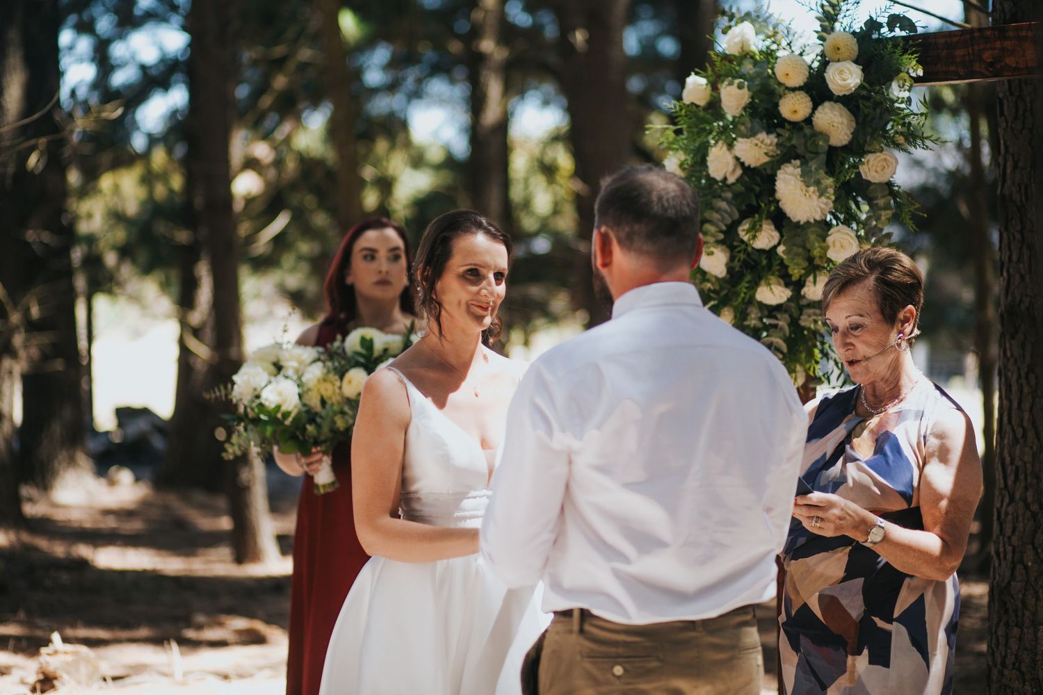 Bride and groom exchange vows beneath floral arch at outdoor forest wedding with bridal party.