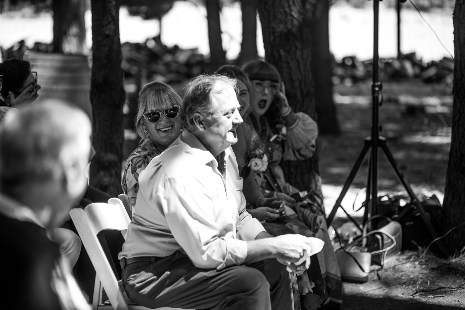 Black and white photo of wedding guests seated outdoors, smiling during forest ceremony.