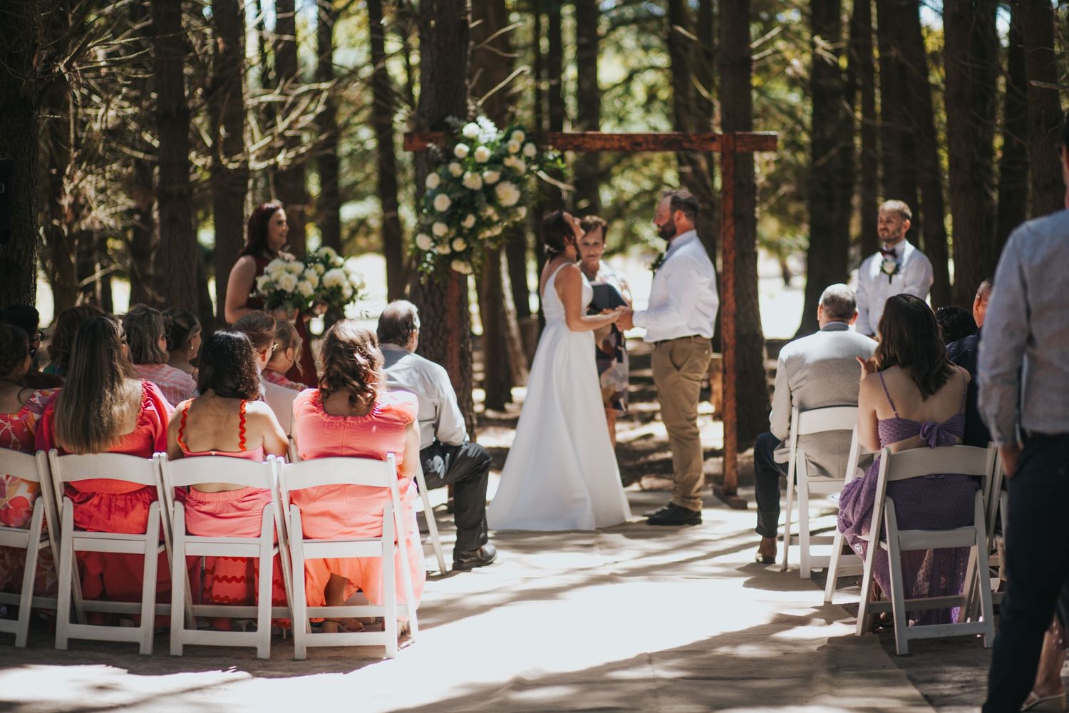 Outdoor forest wedding ceremony with guests seated in white chairs as bride and groom exchange vows at rustic wooden arch.
