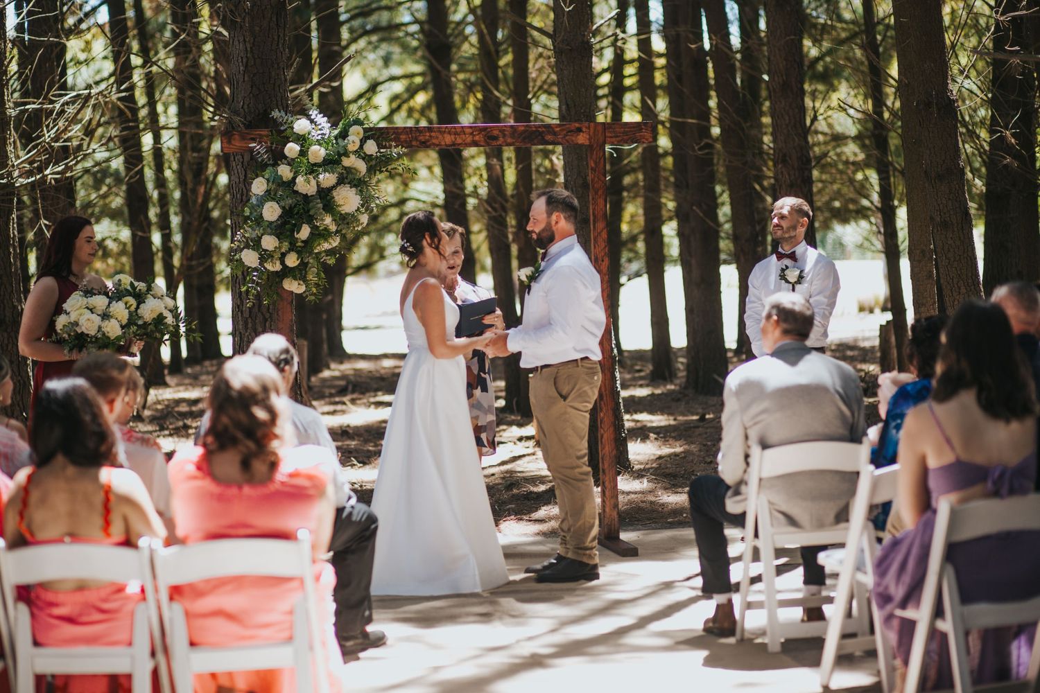Bride and groom face each other during outdoor woodland wedding ceremony with floral arch and seated guests.