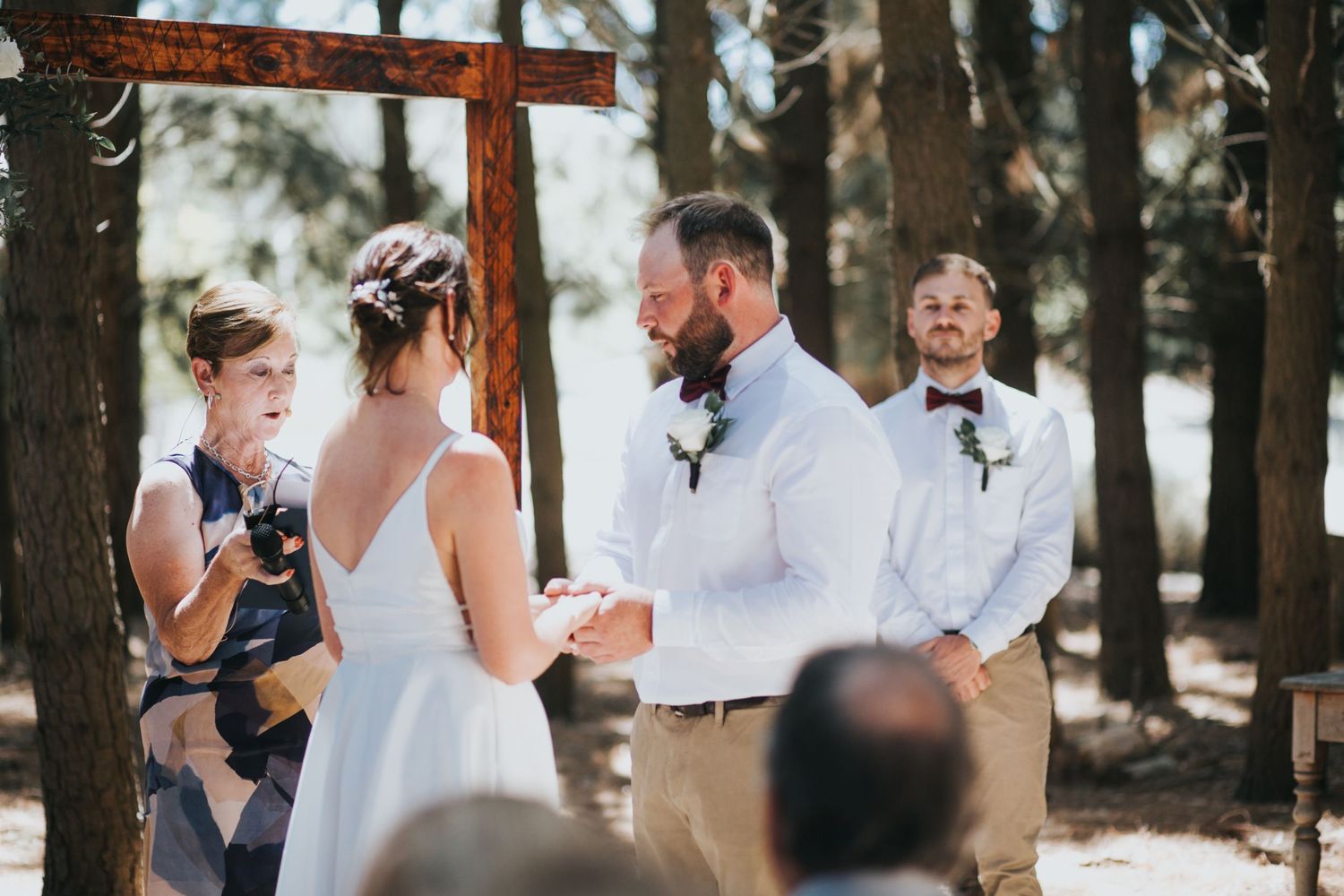 Bride and bearded groom exchange vows at rustic cross altar during sunlit outdoor forest wedding ceremony.