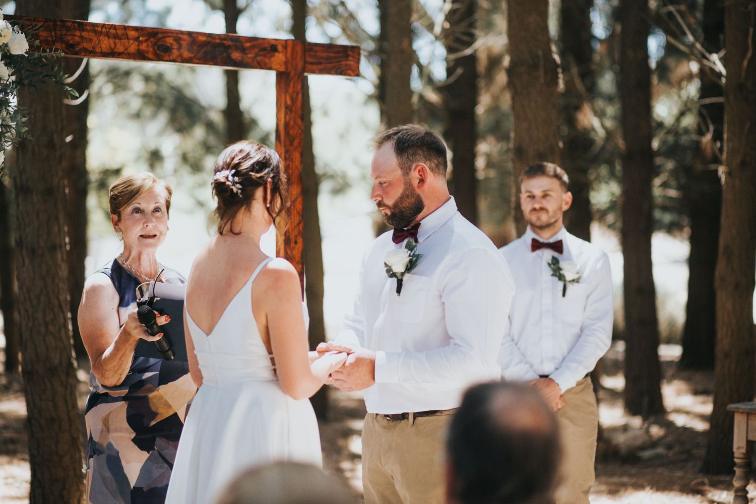 Bride reads vows to bearded groom at rustic wooden cross altar during sunny outdoor forest wedding ceremony.