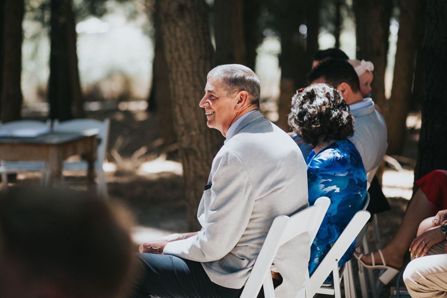 Smiling elderly man in light gray suit seated at outdoor wedding ceremony among guests in a forest setting.