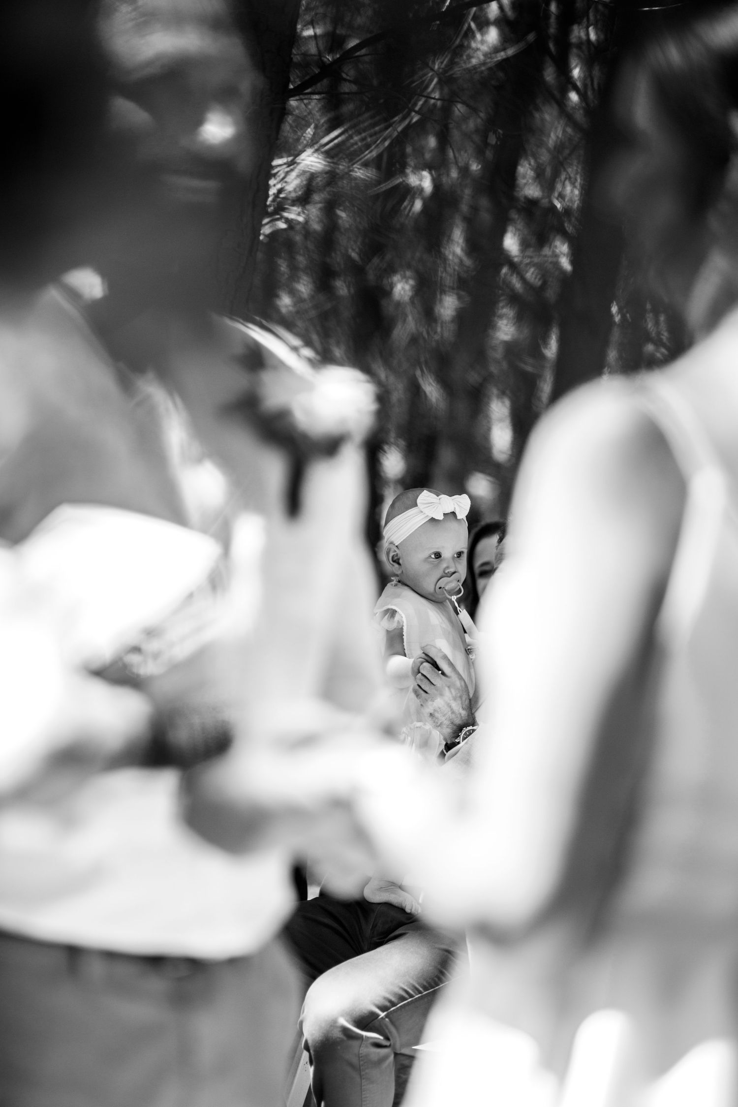 Black and white photo of a baby with bow headband being held by guest during an outdoor wedding ceremony.