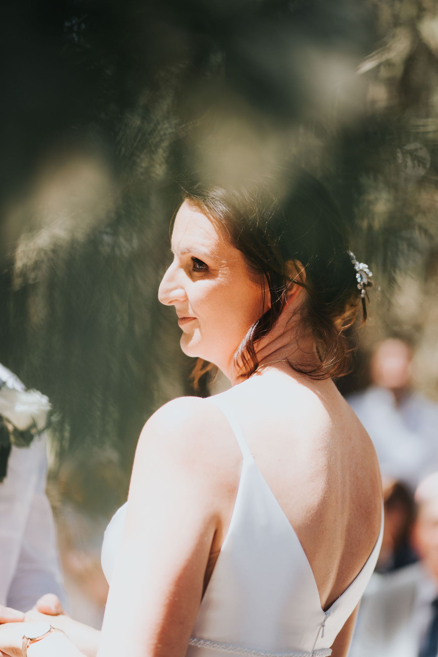 Close-up profile portrait of bride in backless white gown with floral hair accessory at outdoor wedding.