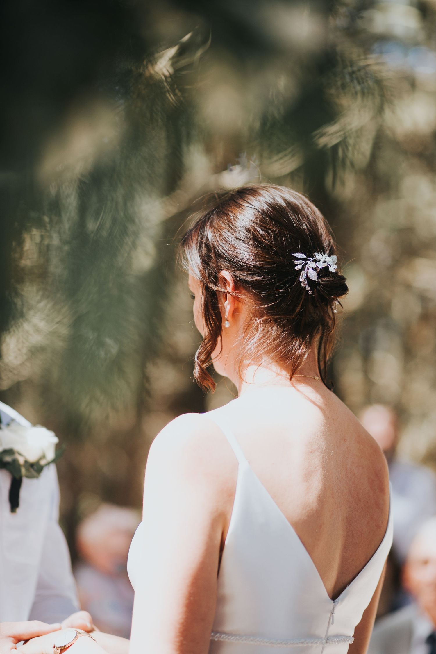 Elegant bride with updo hairstyle adorned with jeweled hair clip in backless white gown at outdoor wedding.