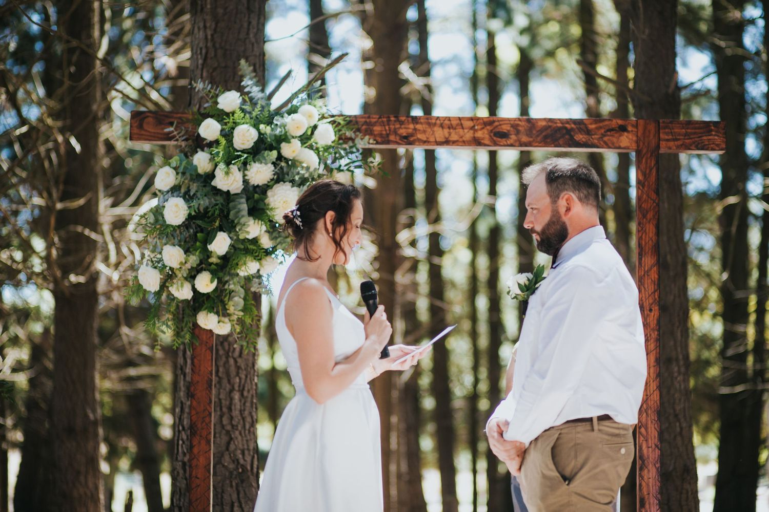 Bride reads personal vows to groom beneath floral-decorated rustic wooden cross arch in sunlit forest ceremony.