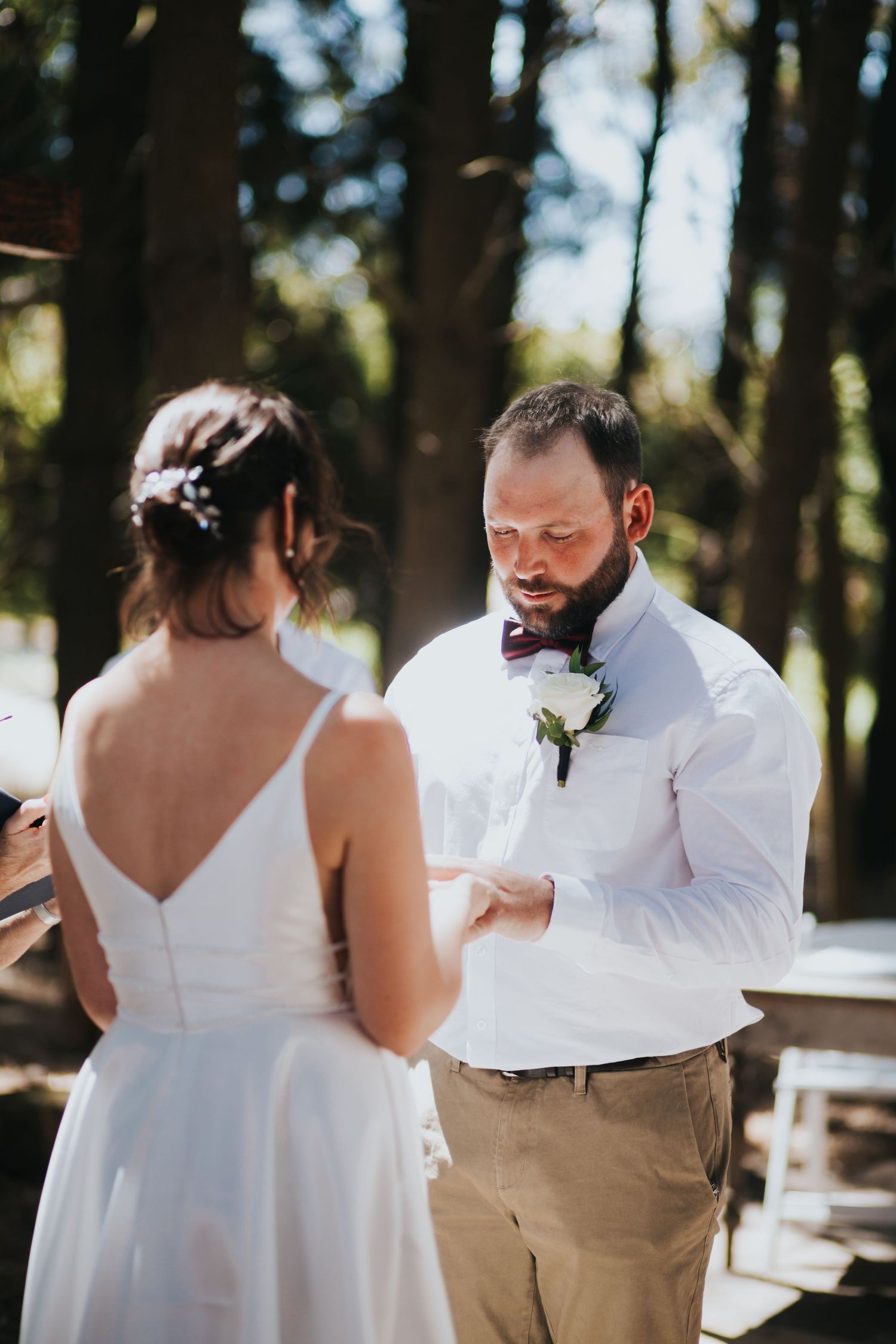 Emotional bearded groom in white shirt and burgundy bow tie listens to bride during outdoor wedding vows.