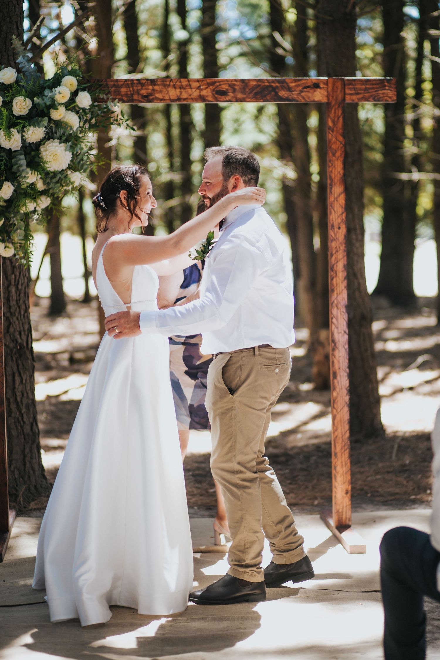 Bride and groom share a joyful first dance under a wooden arch adorned with white flowers in a sunlit forest.