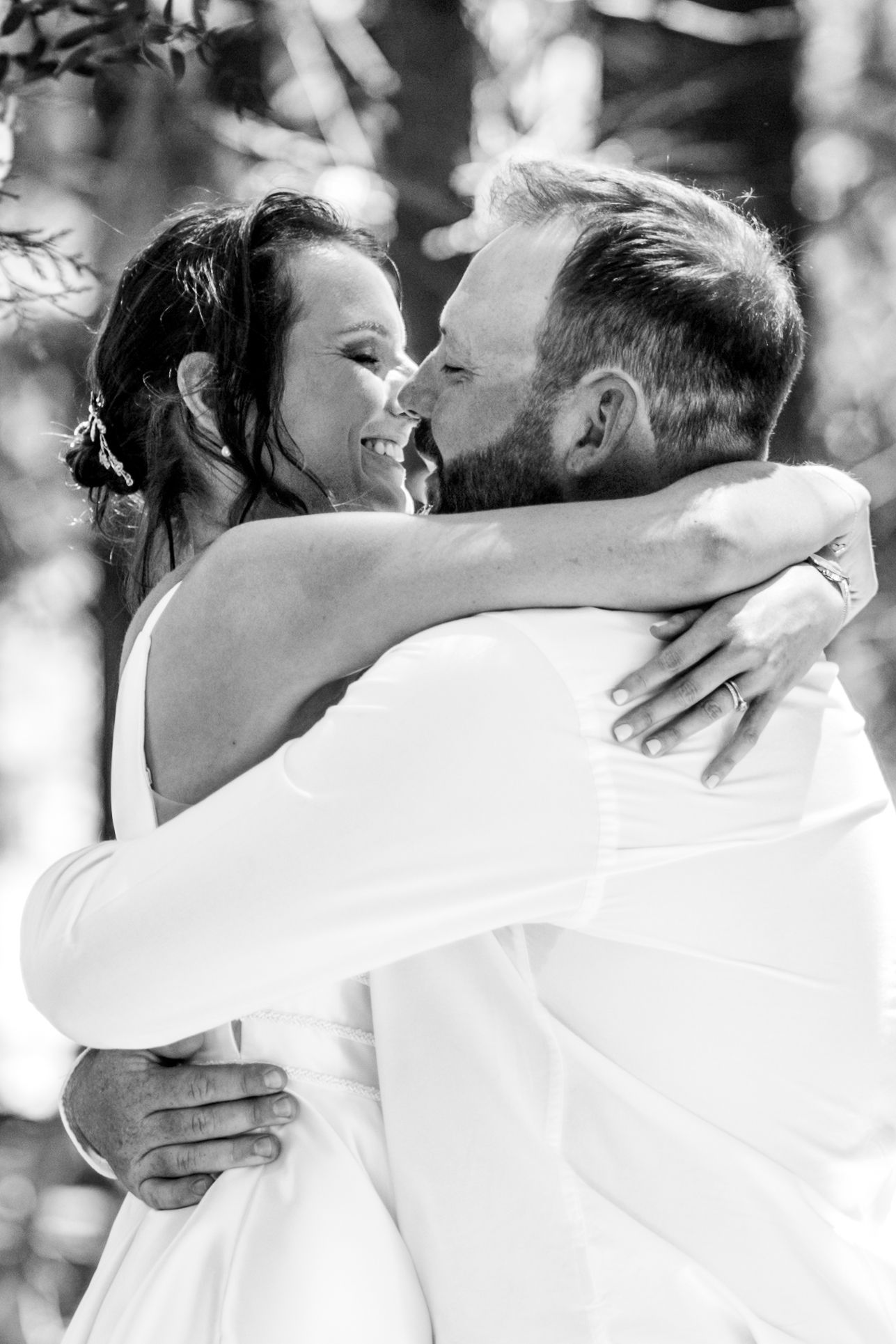 Black and white photo of a smiling bride and groom sharing a tender embrace outdoors on their wedding day.