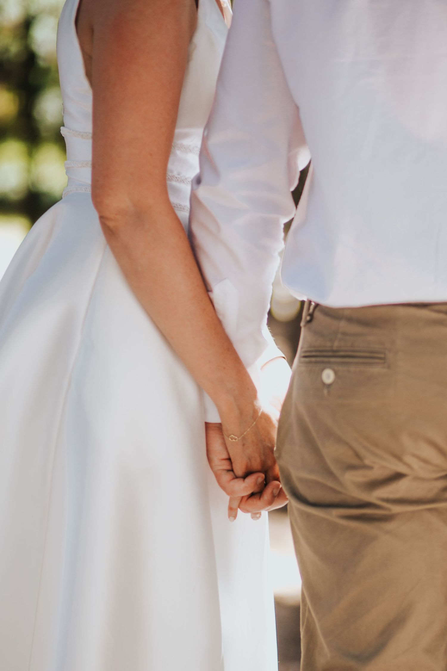 Close-up of a bride in a white gown and groom in khaki pants holding hands during their outdoor wedding.