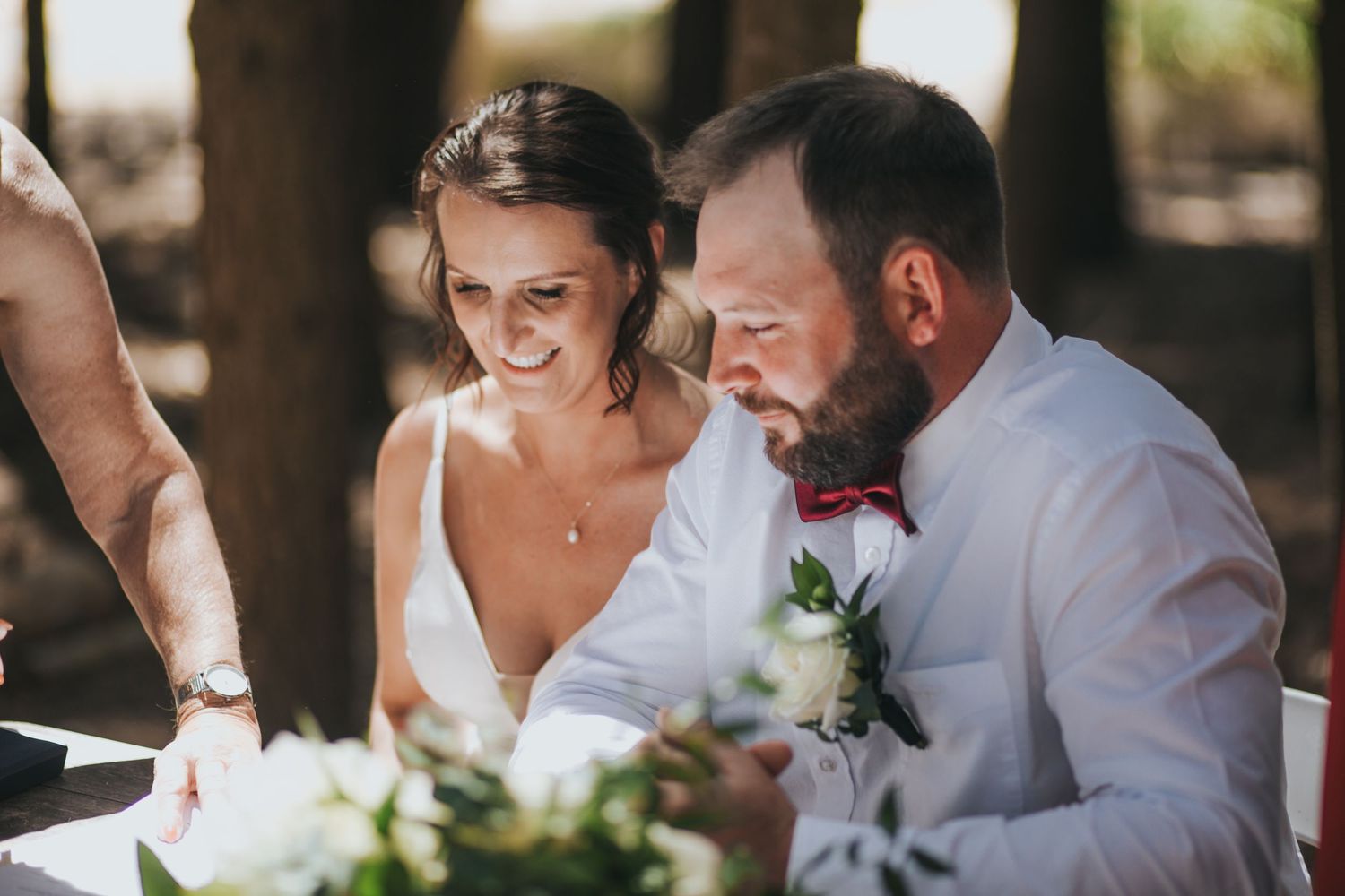 Smiling bride and groom signing their marriage certificate at an outdoor woodland wedding ceremony.