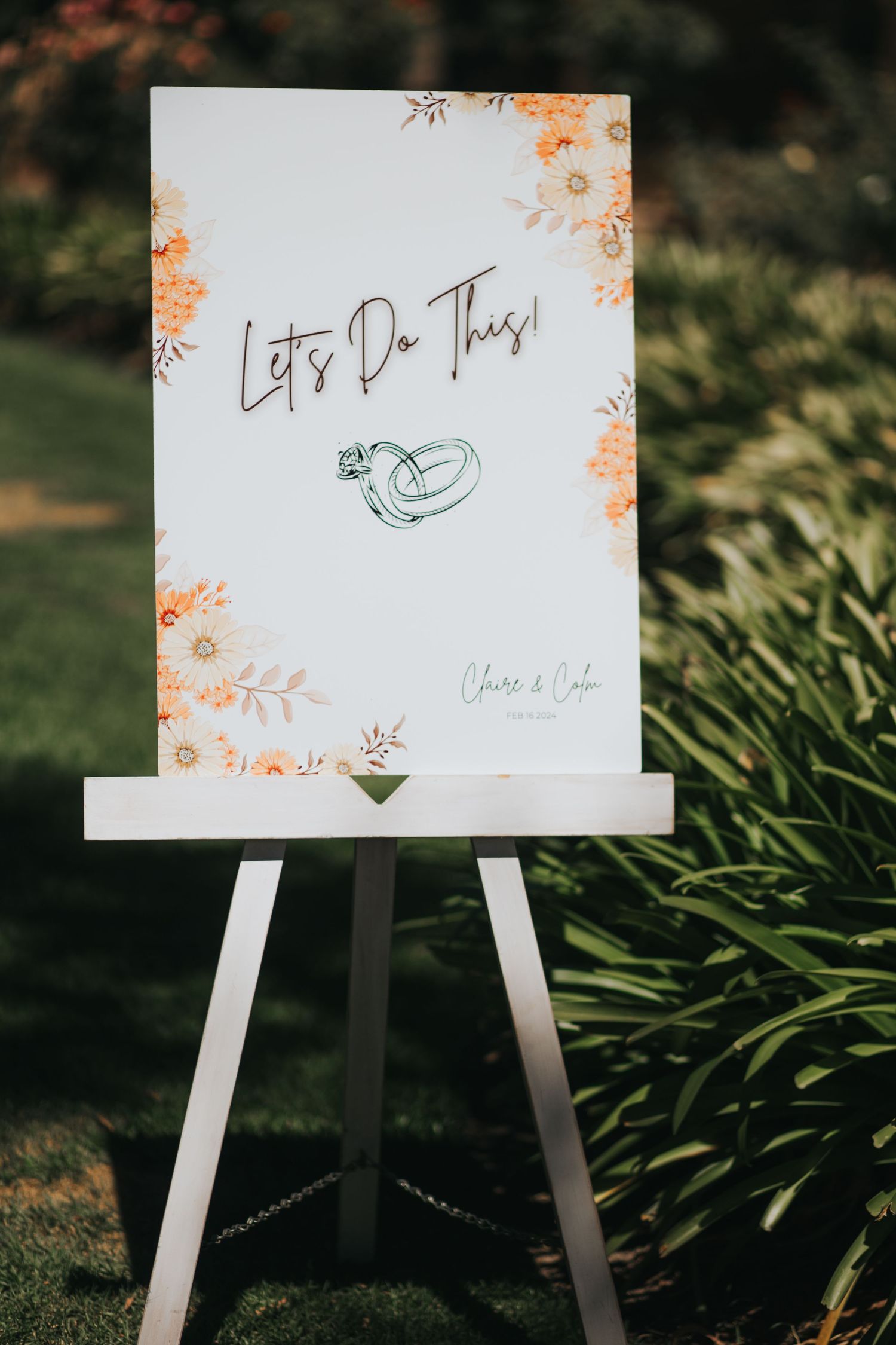 Elegant wedding welcome sign reading Let's Do This with floral details on a white easel outdoors.