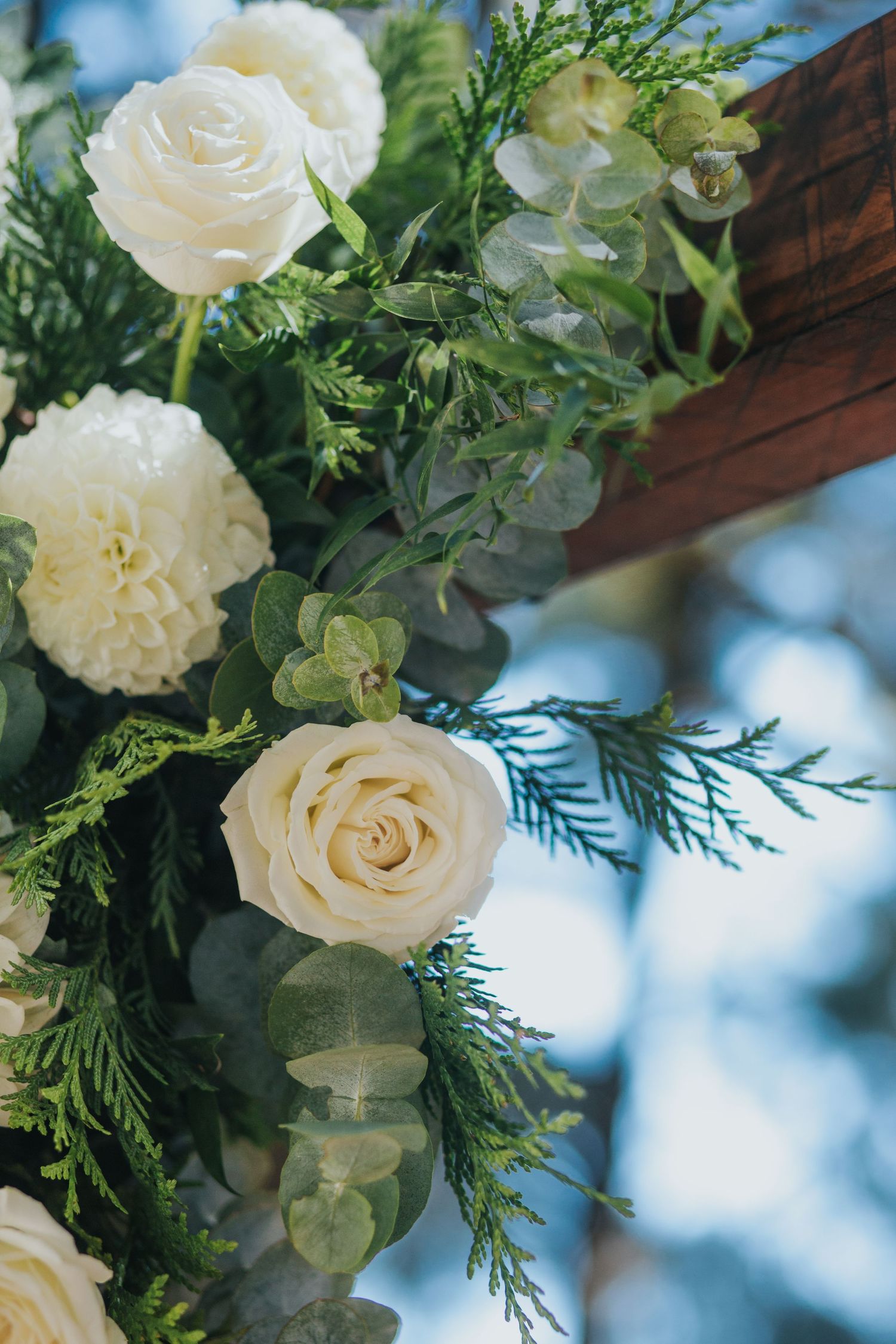 Close-up of white roses and lush greenery adorning a wooden wedding ceremony arch outdoors.