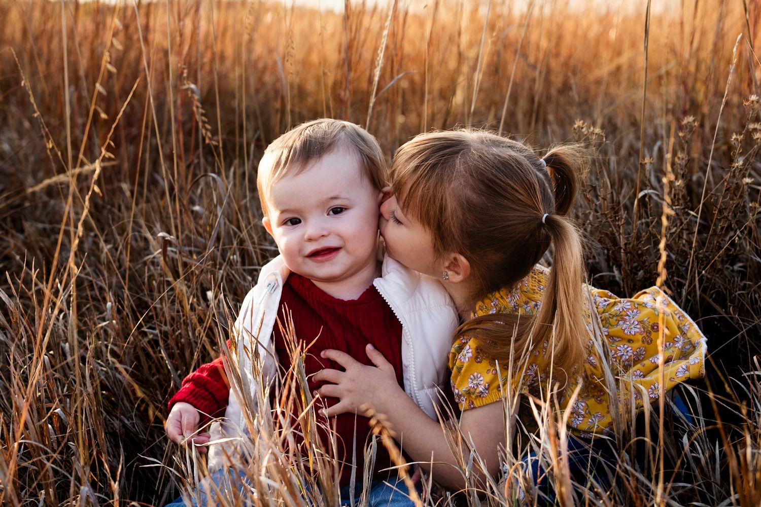 Two children share an affectionate moment in golden autumn grass at sunset.