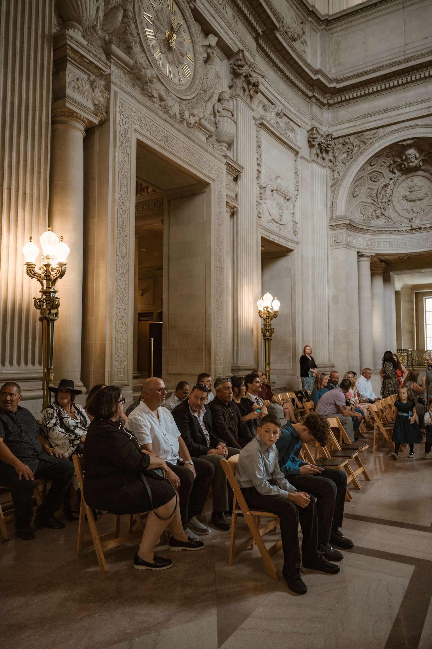 Wedding guests seated in wooden chairs inside an ornate classical building with tall columns and chandeliers.