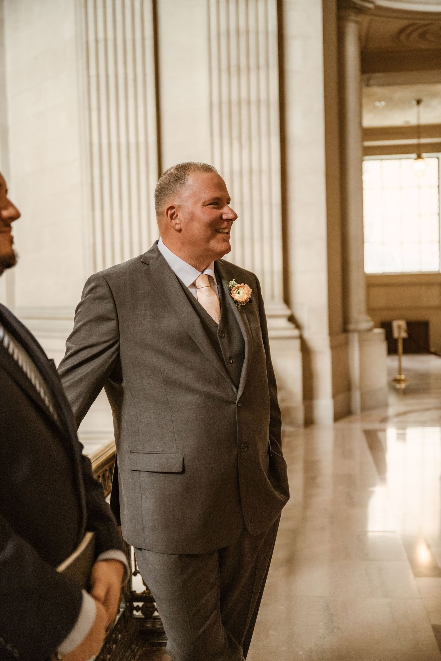 A groom in a grey suit smiles while waiting in an elegant hallway with classical architecture.