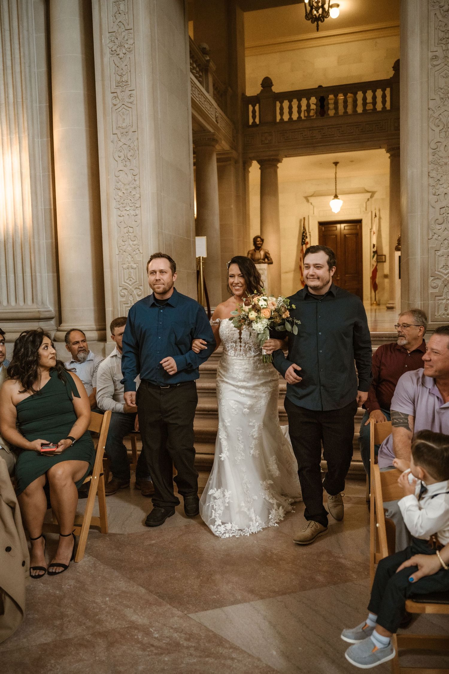 Wedding party members seated in a row along an ornate hallway with classical columns and decorative details.