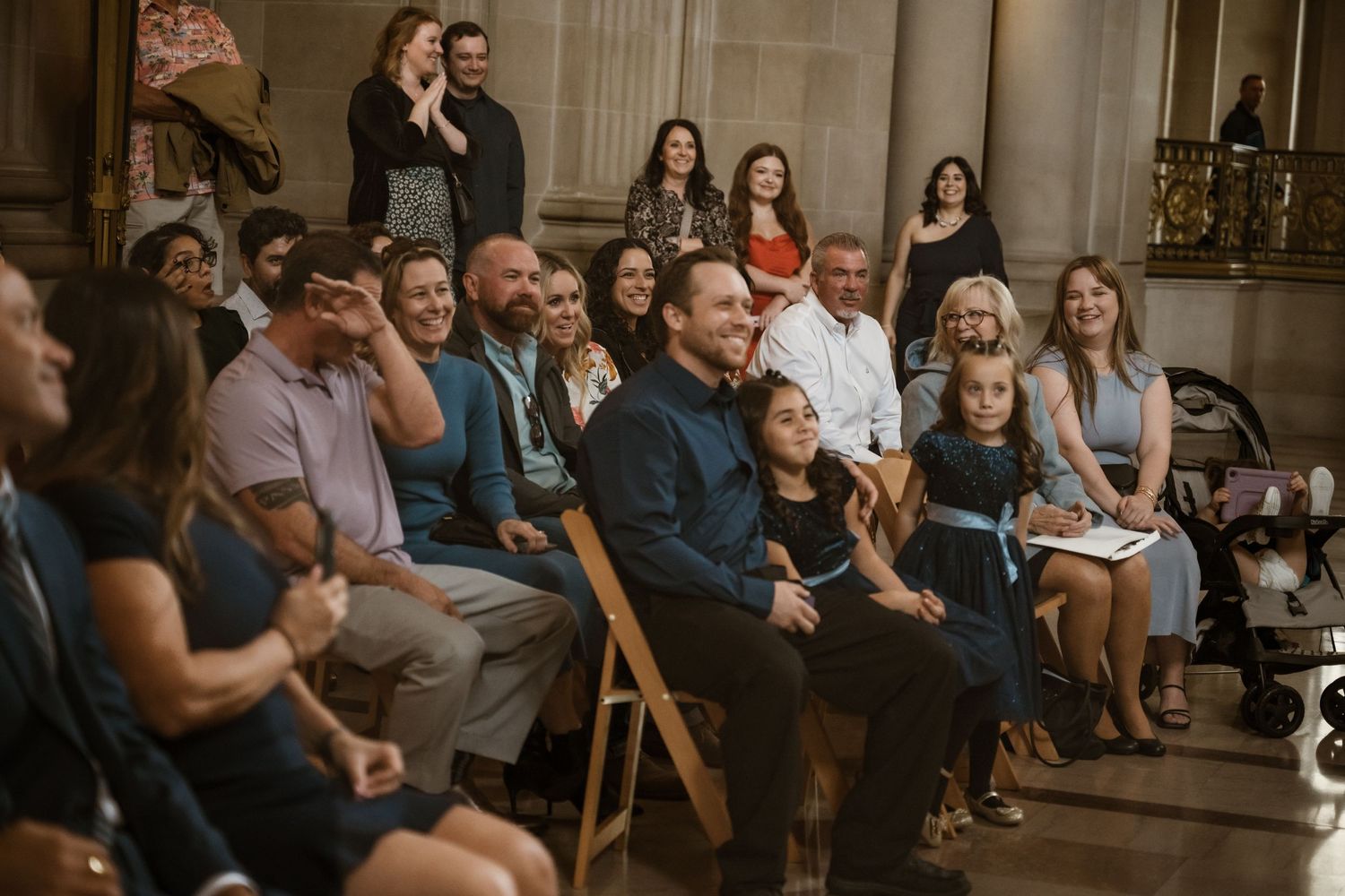 Wedding guests watching ceremony in grand historic venue with ornate columns and decor.