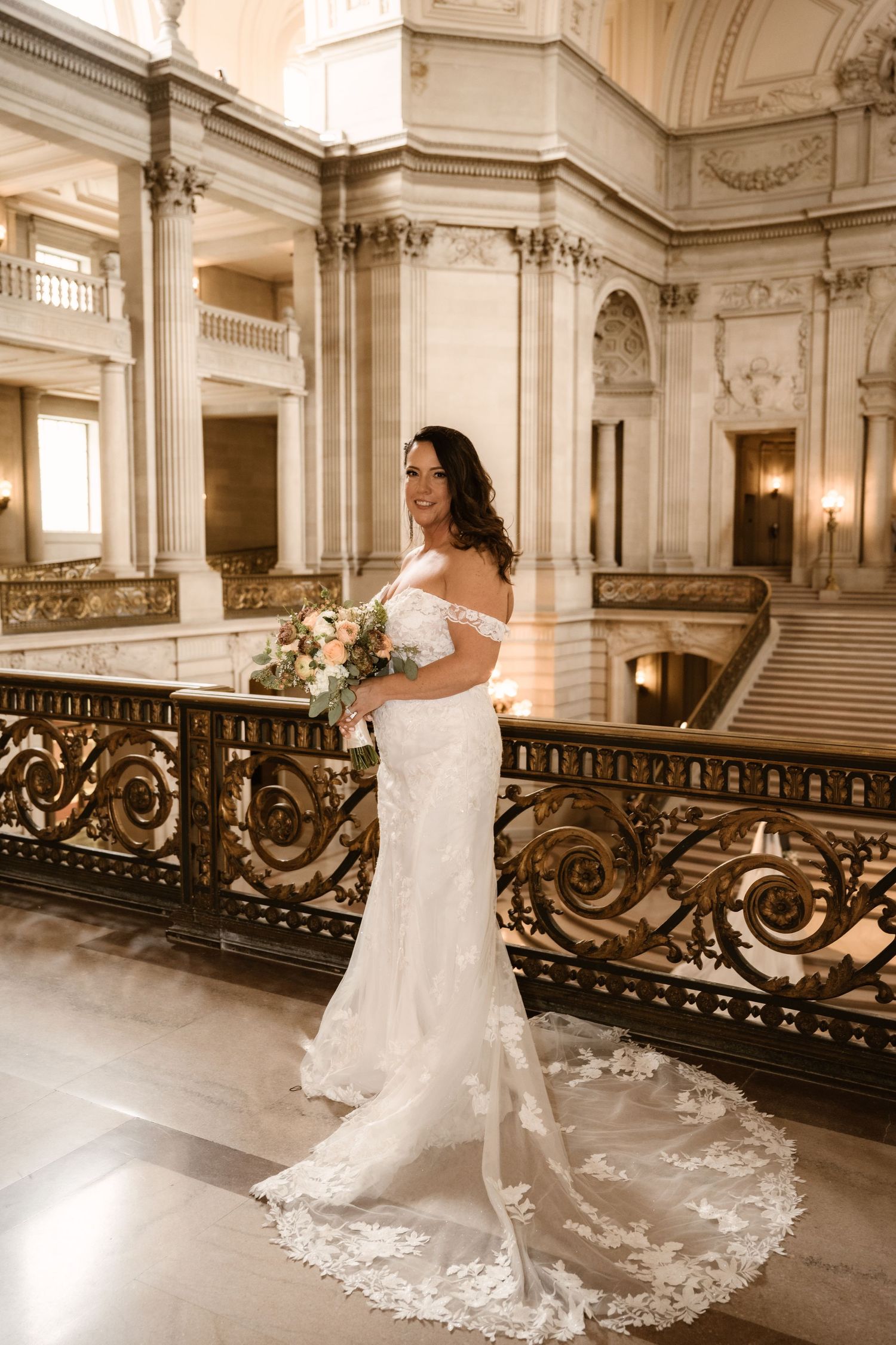Bride in flowing white dress stands alone on elegant marble staircase with ornate railings.