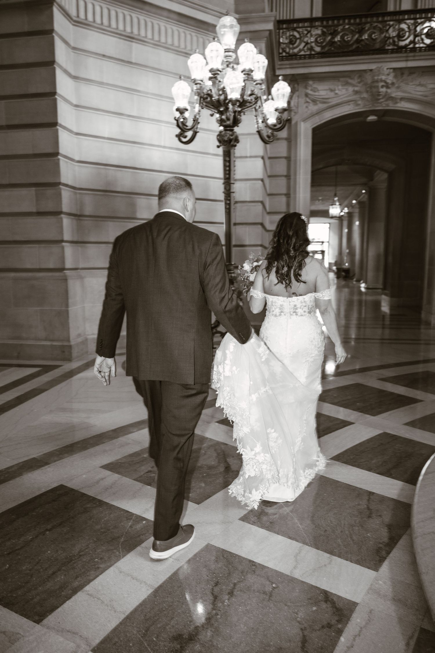 Black and white photo of couple walking through historic building with ornate lighting.