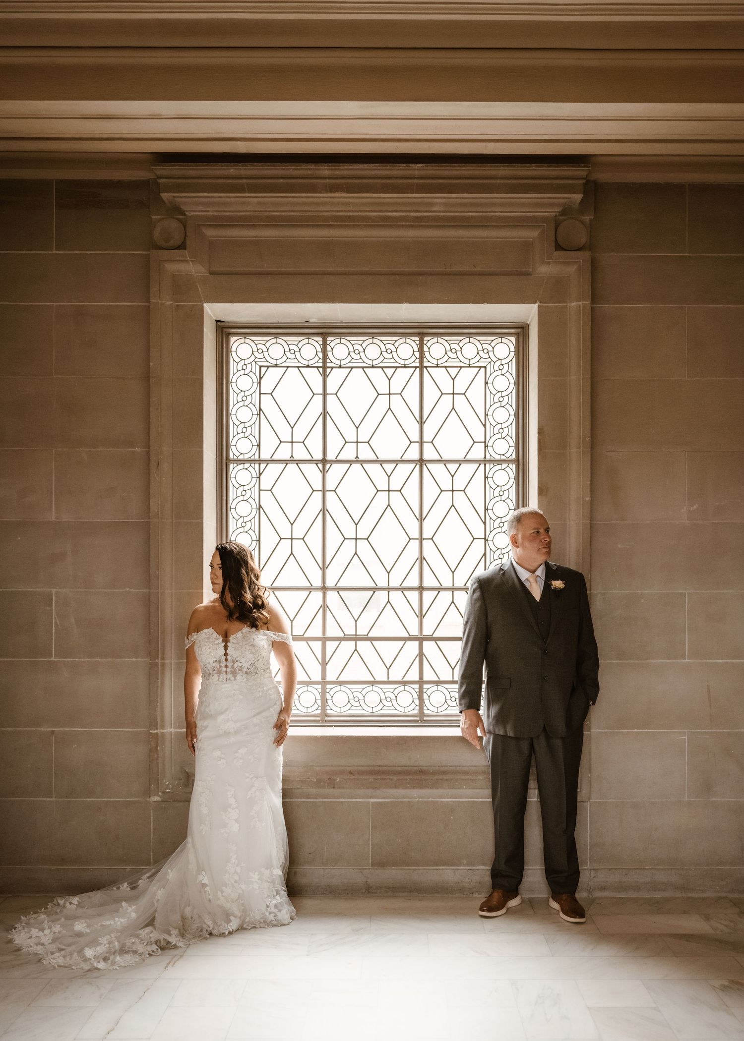 Elegant bride in flowing white gown stands across from groom near ornate window in stone room.