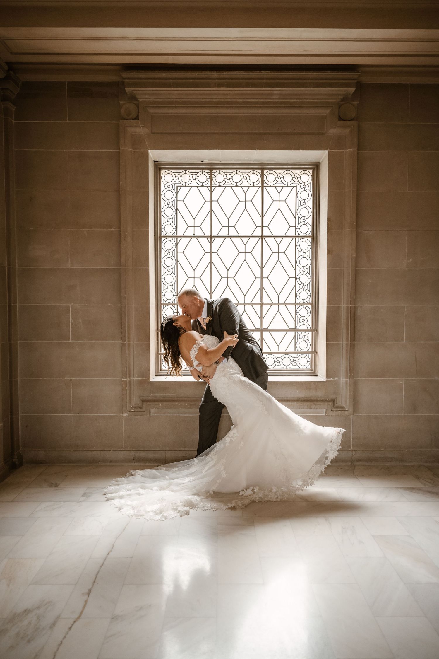 Bride and groom dance gracefully in sunlit room with geometric window pattern.