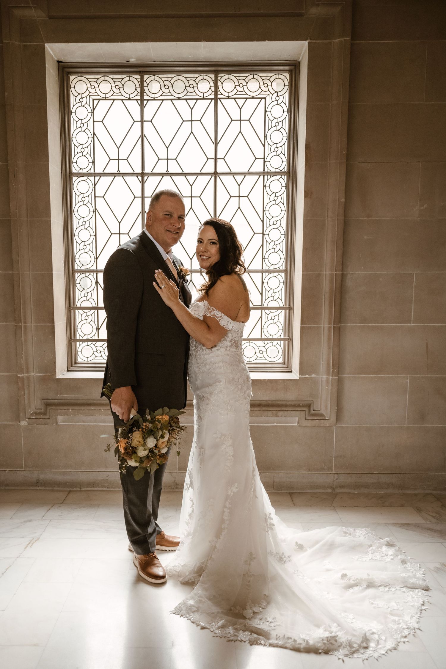 Couple shares intimate wedding moment in front of ornate window in stone-walled room.