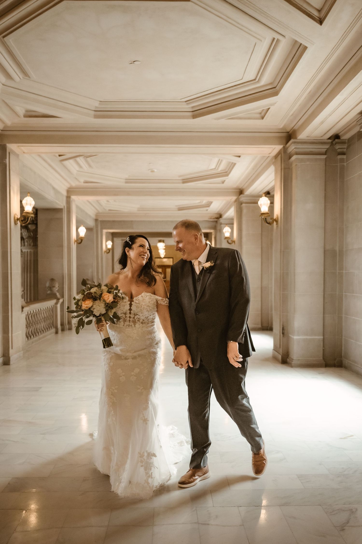 Wedding couple dances through elegant hallway with ornate ceiling and wall sconces.